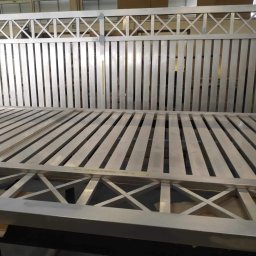 Weld Lab Pro usługi ślusarsko-spawalnicze KAROLINA WOJTECKA - Fragment metalowej, surowej bramy wjazdowej z poprzecznymi szczeblami i dekoracyjnym wzorem krzyży, w hali produkcyjnej.