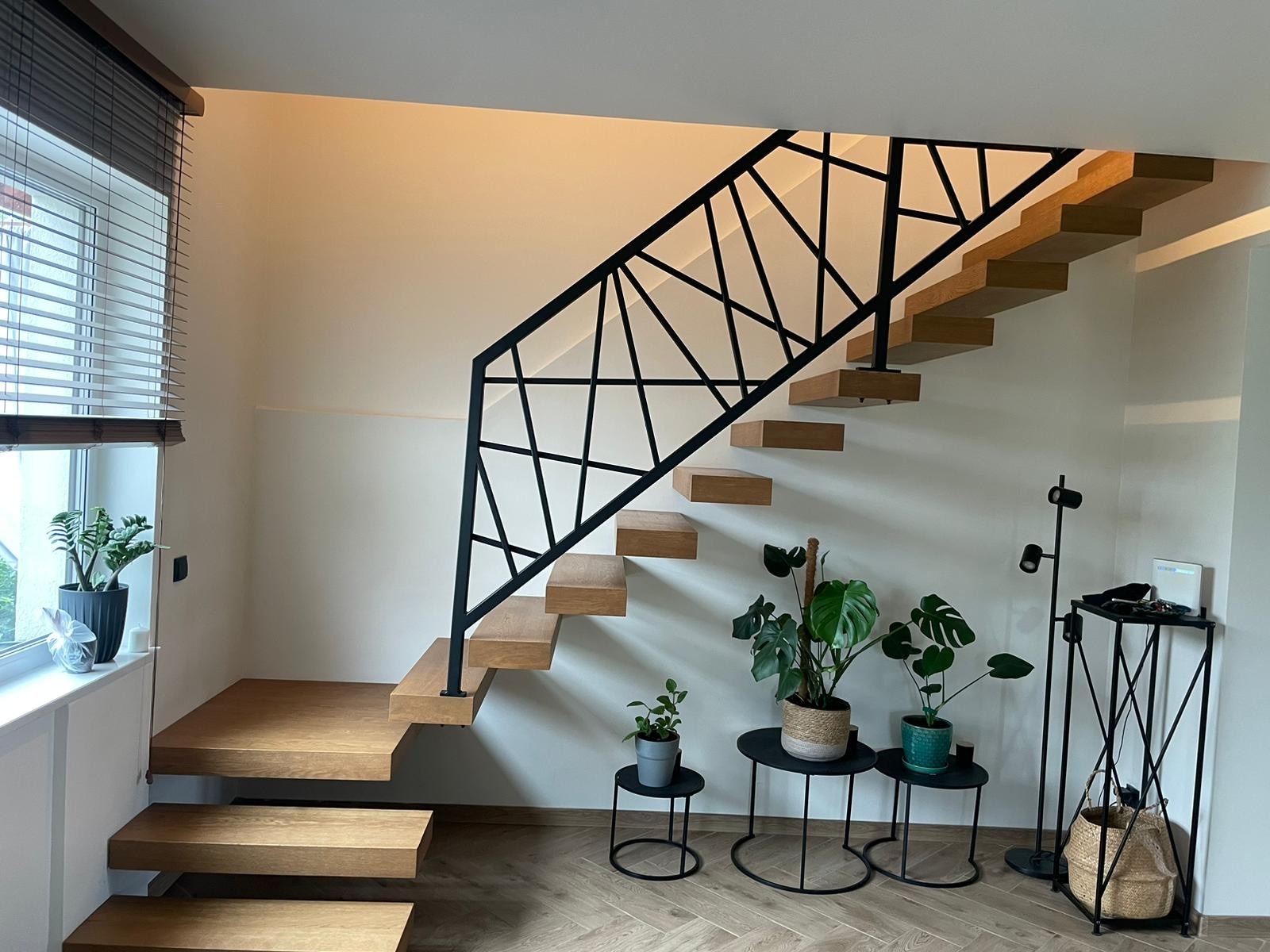 Nowoczesne schody drewniane z czarną, geometryczną balustradą, oświetlone naturalnym światłem z okna, uzupełnione dekoracyjnymi roślinami w doniczkach.