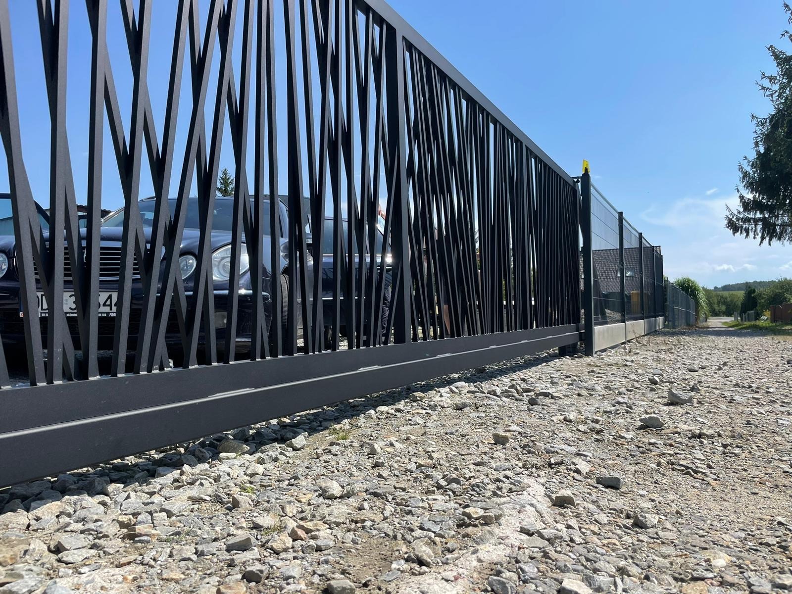 Czarna brama wjazdowa z pionowymi, nieregularnymi prętami, widok z dołu na żwirową nawierzchnię, w tle zaparkowany ciemny samochód i błękitne niebo.