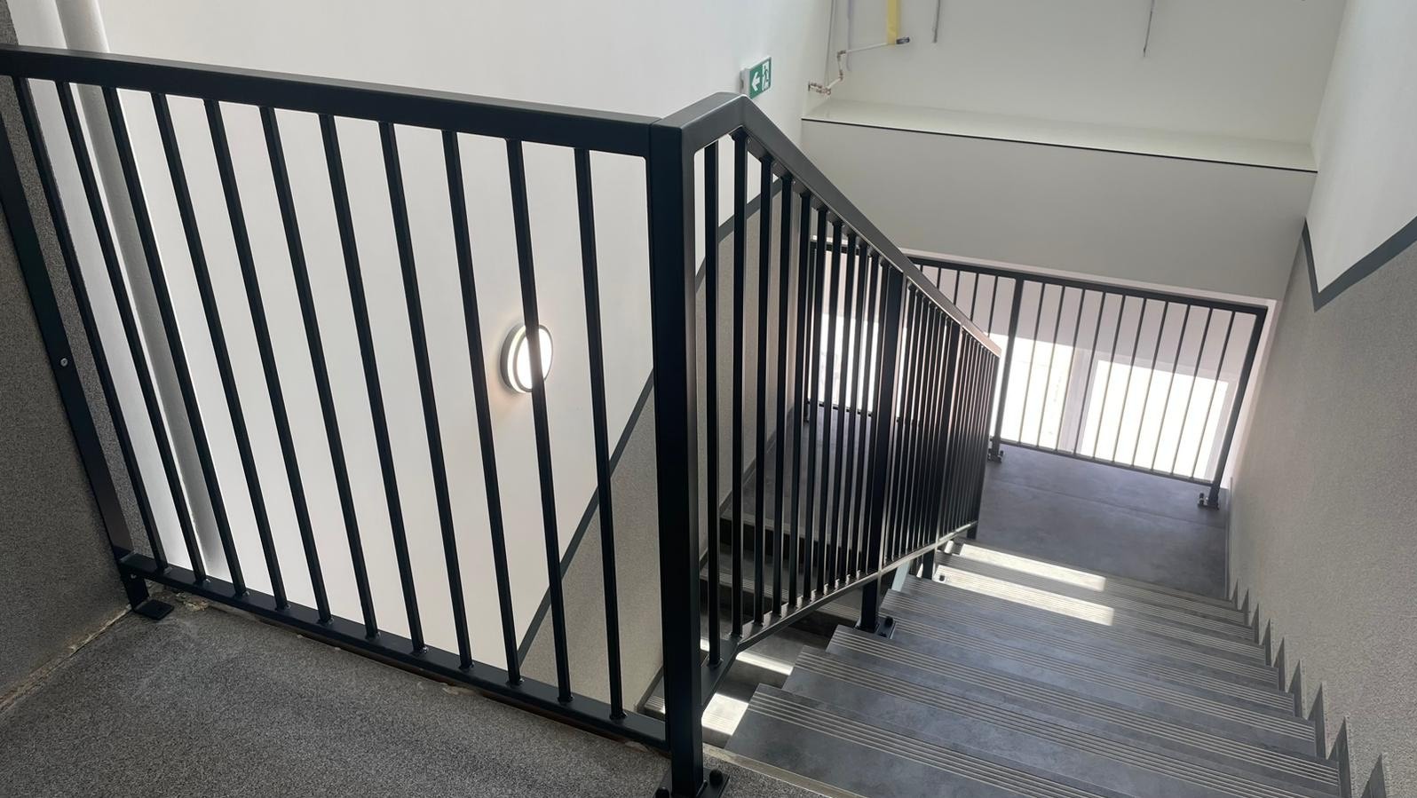 Klatka schodowa z czarną, metalową balustradą z pionowymi prętami; widoczne stopnie z antypoślizgowymi paskami i lampa ścienna.