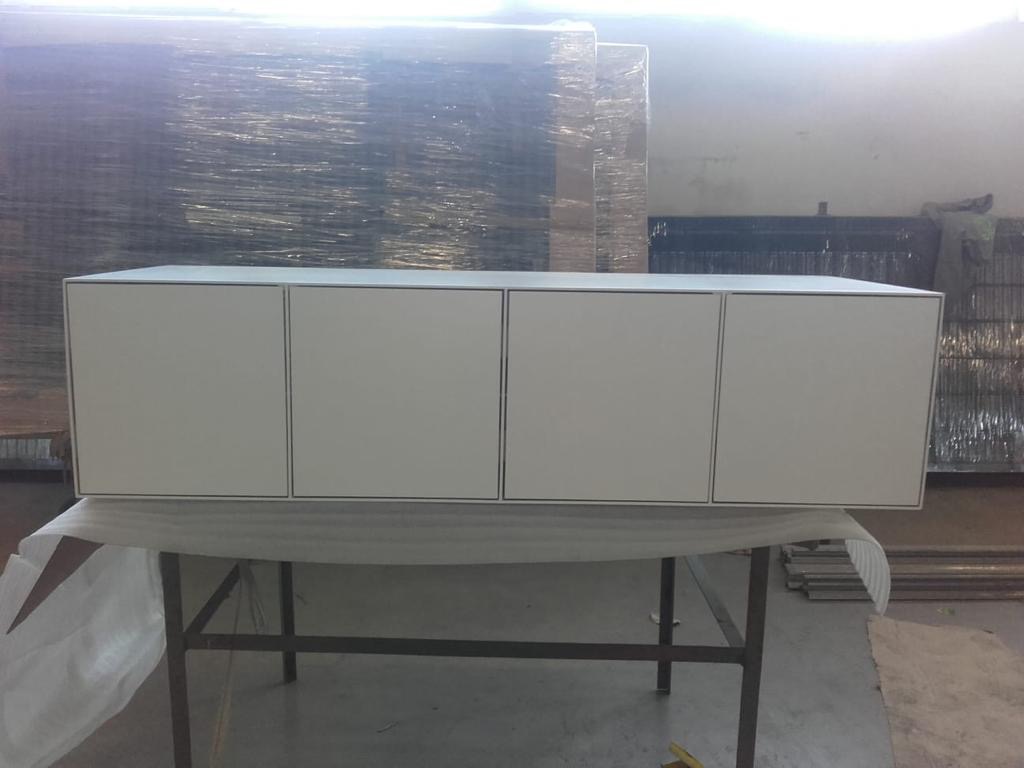 Biała szafka wisząca z czterema drzwiczkami, umieszczona na metalowym stole w warsztacie, z widocznymi materiałami budowlanymi w tle.