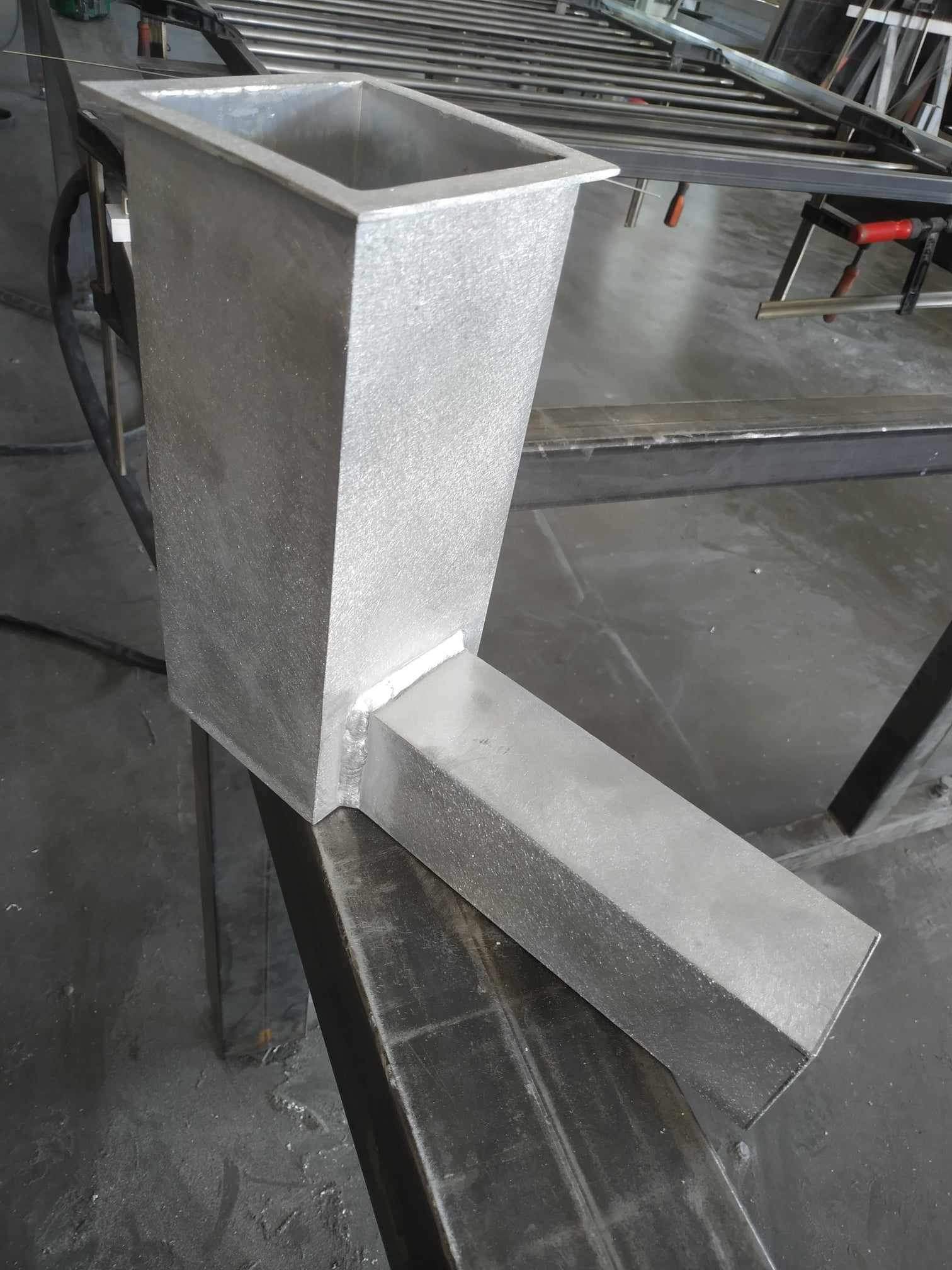 Metalowy pojemnik z prostokątnym otworem wlotowym i wylotowym, spawany z blachy, umieszczony na metalowej konstrukcji w warsztacie.