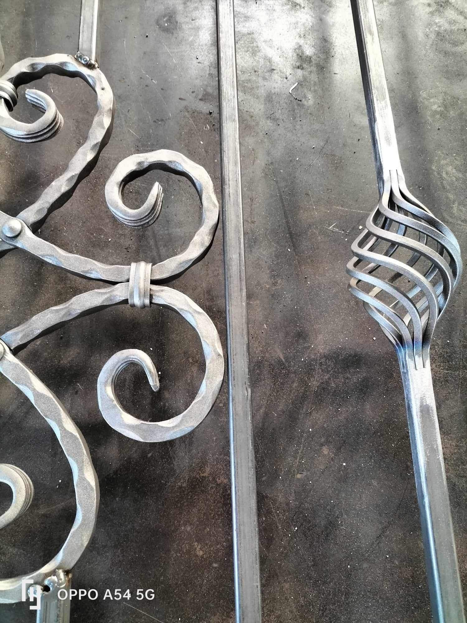 Elementy dekoracyjne z metalu, prawdopodobnie fragment balustrady lub bramy, z widocznymi spawami i spiralnym zdobieniem, leżące na roboczym, metalowym stole.