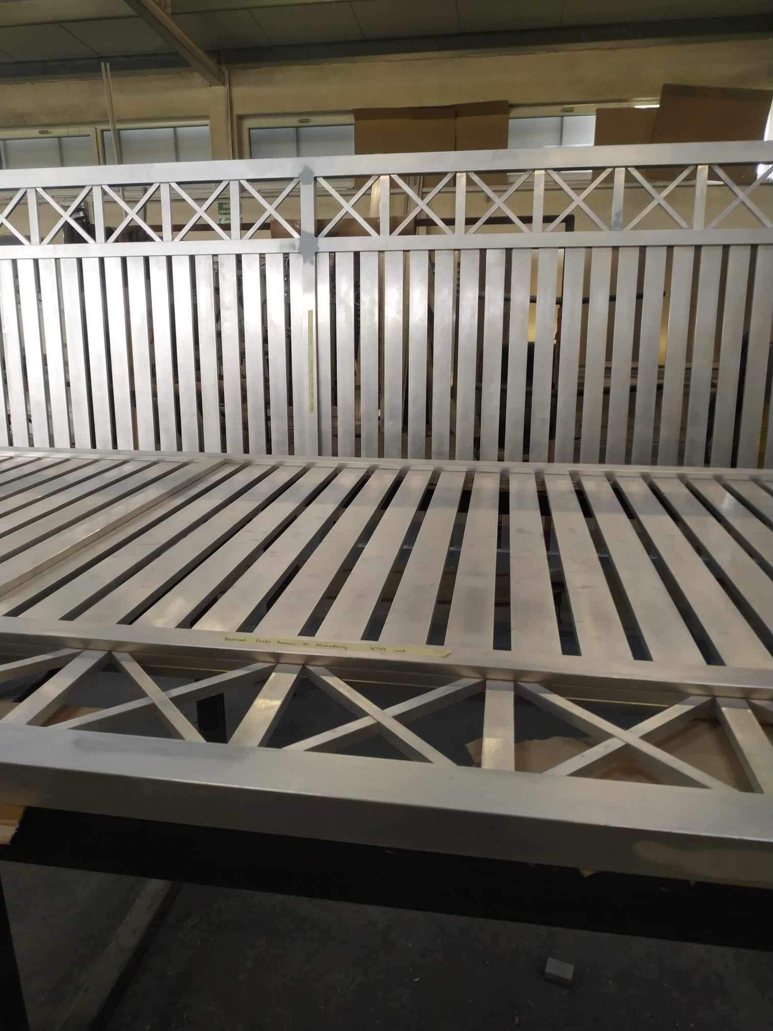 Fragment metalowej, surowej bramy wjazdowej z poprzecznymi szczeblami i dekoracyjnym wzorem krzyży, w hali produkcyjnej.