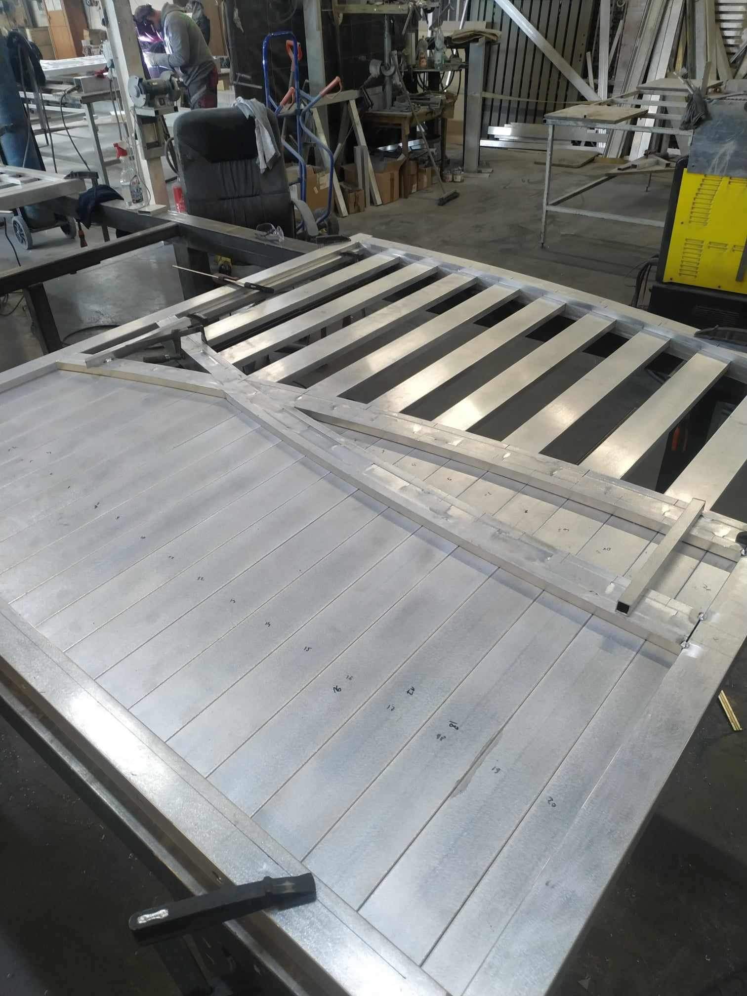 Wnętrze warsztatu z widoczną konstrukcją metalową bramy wjazdowej w trakcie spawania, z numerami naniesionymi na elementach, spawarka w tle.