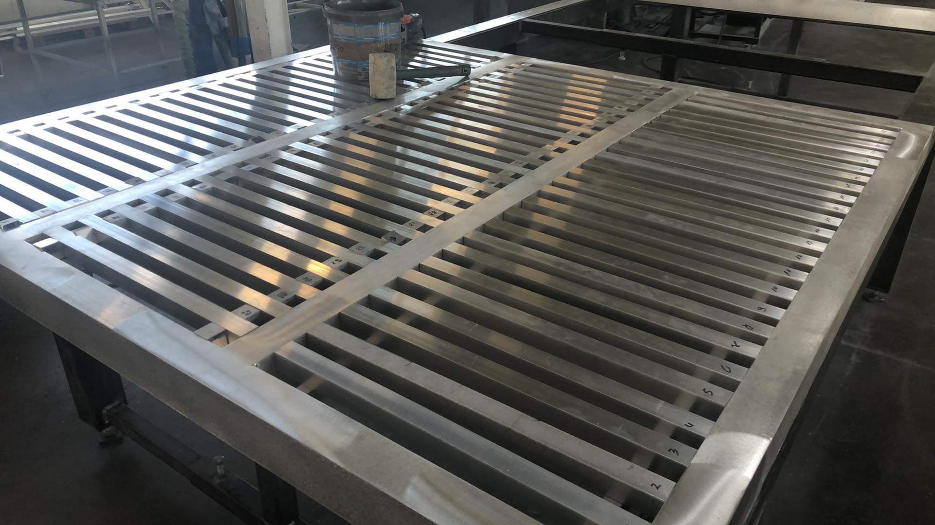 Metalowa konstrukcja bramy w trakcie produkcji, z numeracją poszczególnych elementów, widoczna puszka z farbą i narzędzia na blacie roboczym.