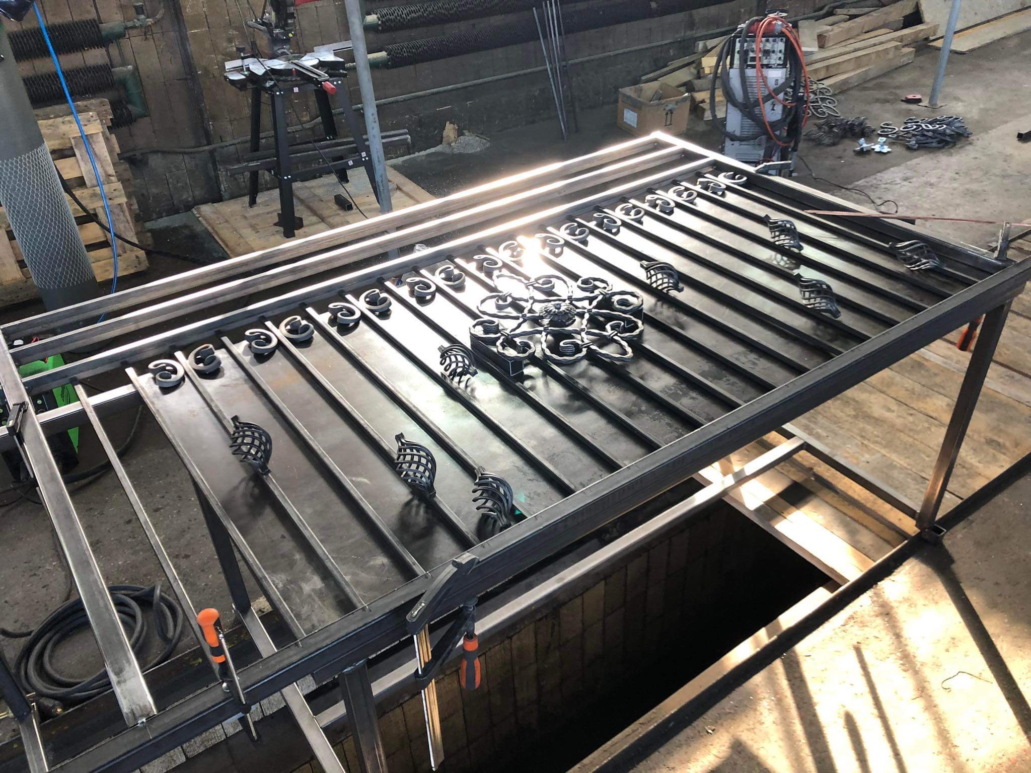 Metalowa konstrukcja bramy z dekoracyjnymi elementami w warsztacie, widoczne narzędzia i spawarka w tle.