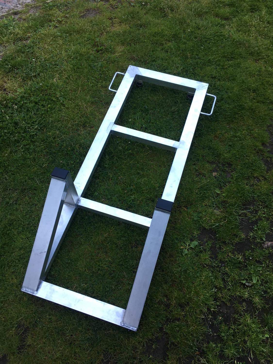 Surowa, metalowa konstrukcja z uchwytami, prawdopodobnie podstawa pod urządzenie, leżąca na trawie.