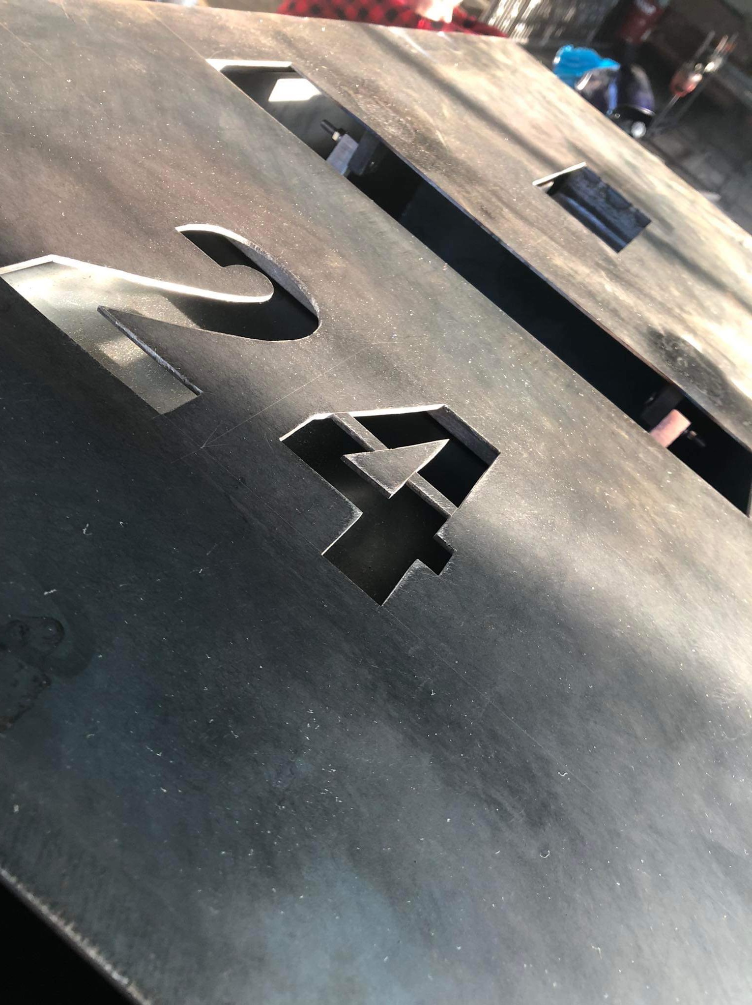 Cięta laserowo, surowa, metalowa płyta z wyciętymi numerami '24', widoczne ślady po obróbce, jasne światło padające z góry.