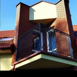 Balkon z kutą, czarną balustradą o dekoracyjnych, giętych elementach, zamontowany na ceglanym budynku z czerwoną dachówką, widok z dołu na tle błękitnego nieba.