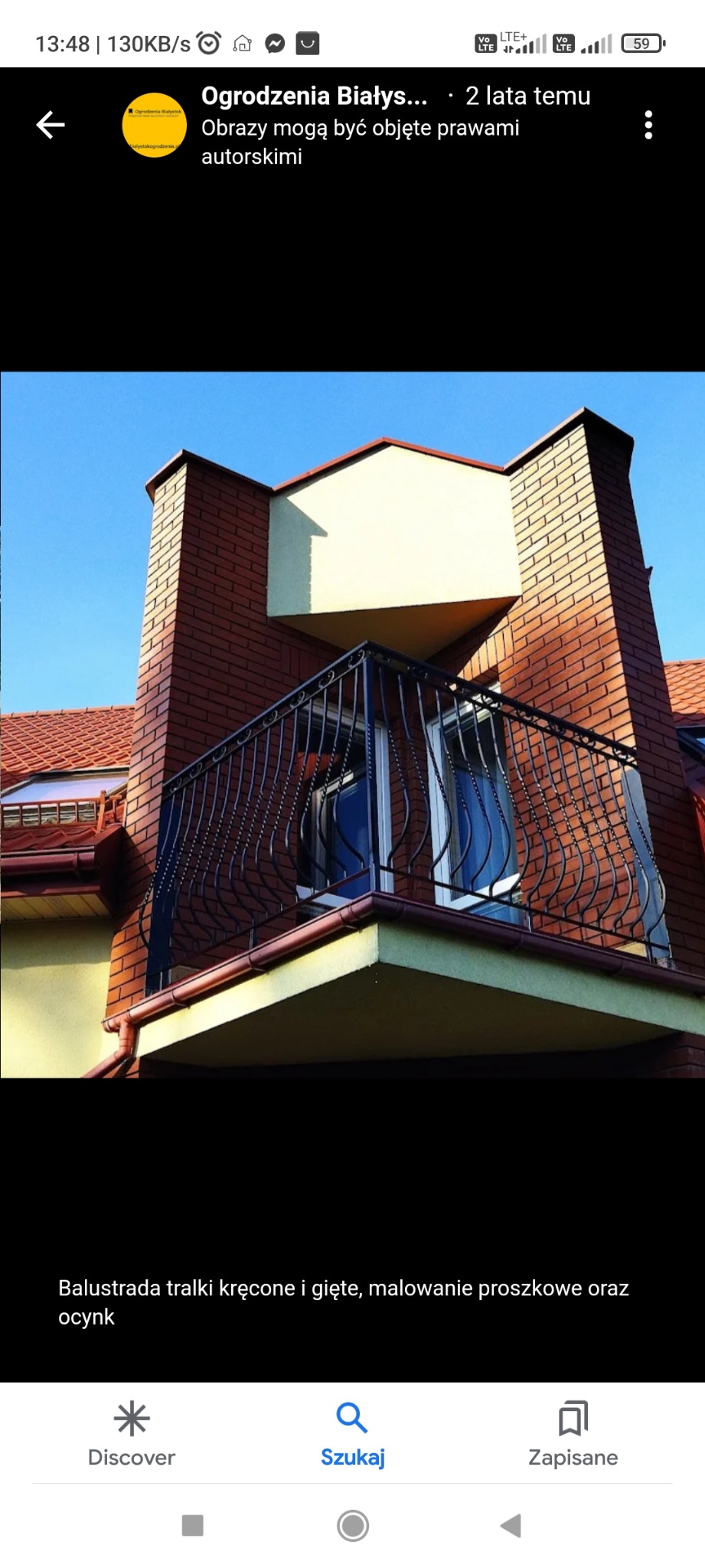 Balkon z kutą, czarną balustradą o dekoracyjnych, giętych elementach, zamontowany na ceglanym budynku z czerwoną dachówką, widok z dołu na tle błękitnego nieba.