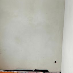 BoTom Tomasz Bogusz - Świeżo położona, jasnoszara gładź na ścianie z widocznym otworem na gniazdko elektryczne i zabezpieczeniem podłogi folią malarską. Minimalistyczne ujęcie.