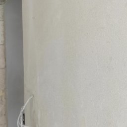 BoTom Tomasz Bogusz - Gładka, jasna ściana po nałożeniu gładzi, z widocznym fragmentem surowego muru i wystającym przewodem elektrycznym. Delikatna faktura powierzchni.