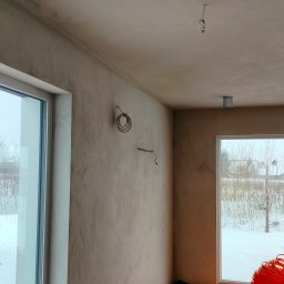 BoTom Tomasz Bogusz - Wnętrze w stanie surowym: ściany pokryte świeżym tynkiem, widoczne przewody elektryczne, okna z widokiem na zimowy krajobraz. Minimalistyczne oświetlenie sufitowe.