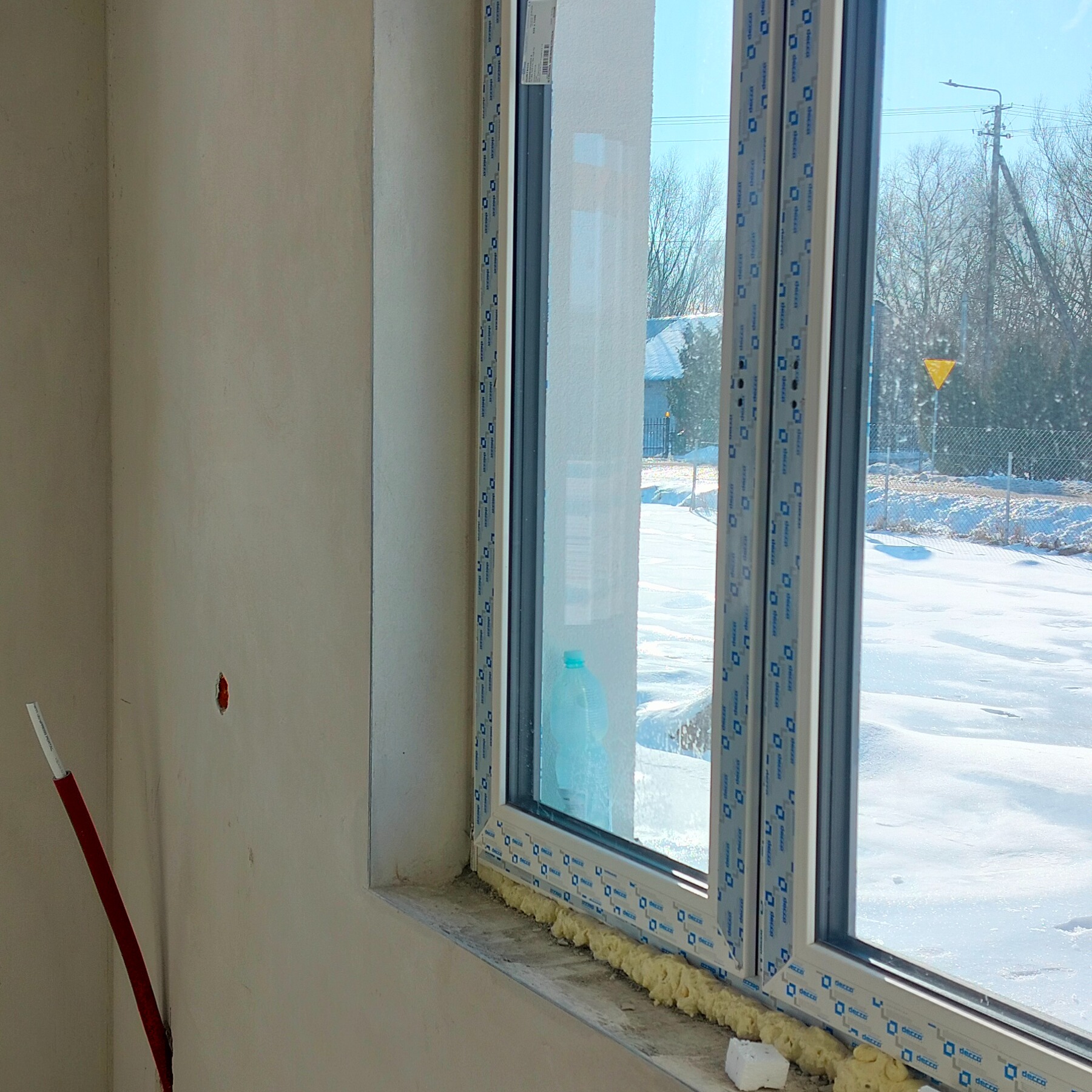 Świeżo osadzone okno PCV z pianką montażową, widok na zimowy krajobraz. Na ścianie widoczne ślady po instalacji elektrycznej. W tle dom i drzewa.