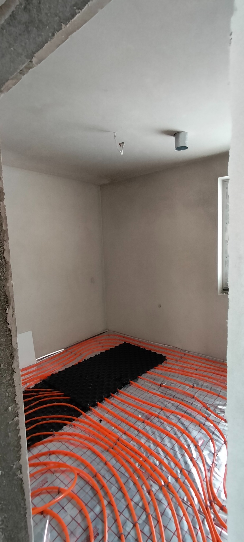 Pomieszczenie w trakcie prac wykończeniowych: widoczne rury ogrzewania podłogowego w kolorze pomarańczowym, częściowo przykryte czarną matą izolacyjną, ściany otynkowane.