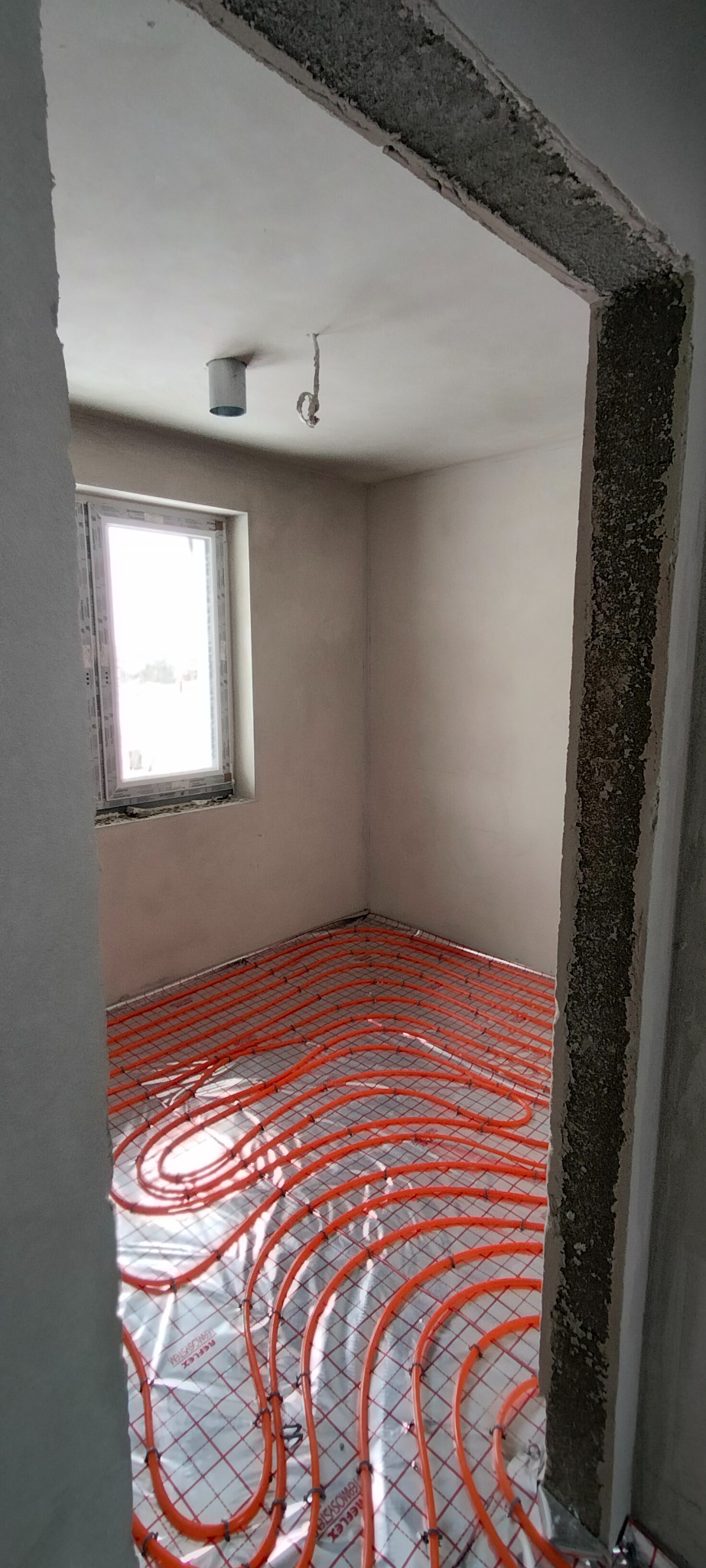 Pomieszczenie w trakcie prac wykończeniowych z widoczną instalacją ogrzewania podłogowego, świeżo położonymi tynkami i surowym wykończeniem ościeżnicy.
