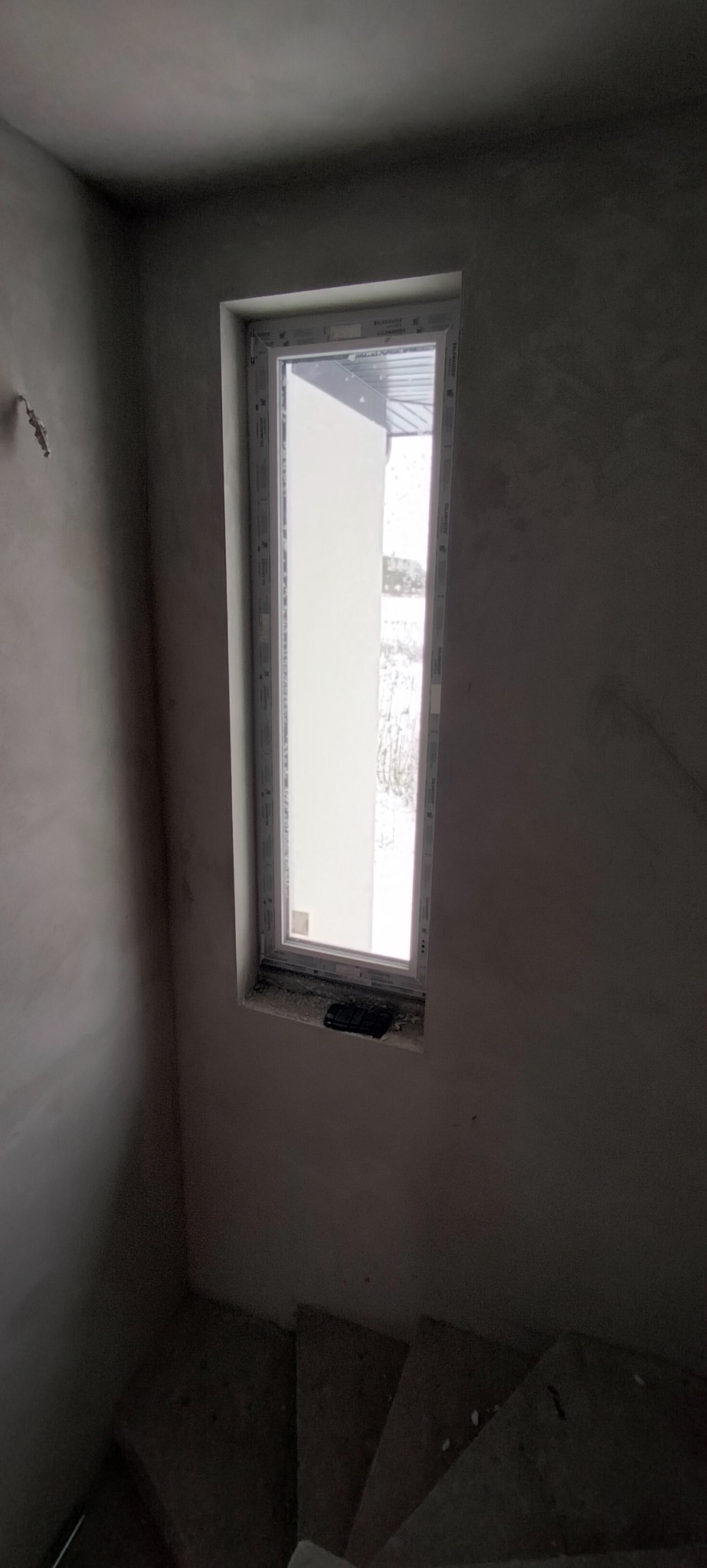 Surowe ściany klatki schodowej po nałożeniu tynku, z widocznym oknem. W tle widoczny zarys budynku. Minimalistyczne ujęcie wnętrza w trakcie prac wykończeniowych.