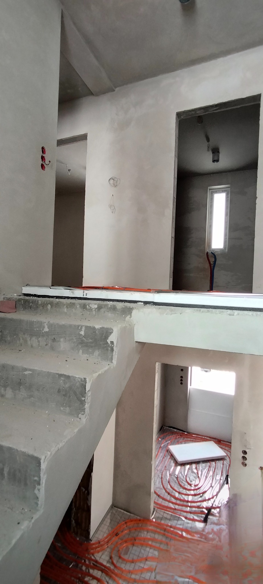 Wnętrze w trakcie wykończenia: surowe schody, ściany po szpachlowaniu, instalacja ogrzewania podłogowego z rurkami w kolorze pomarańczowym.