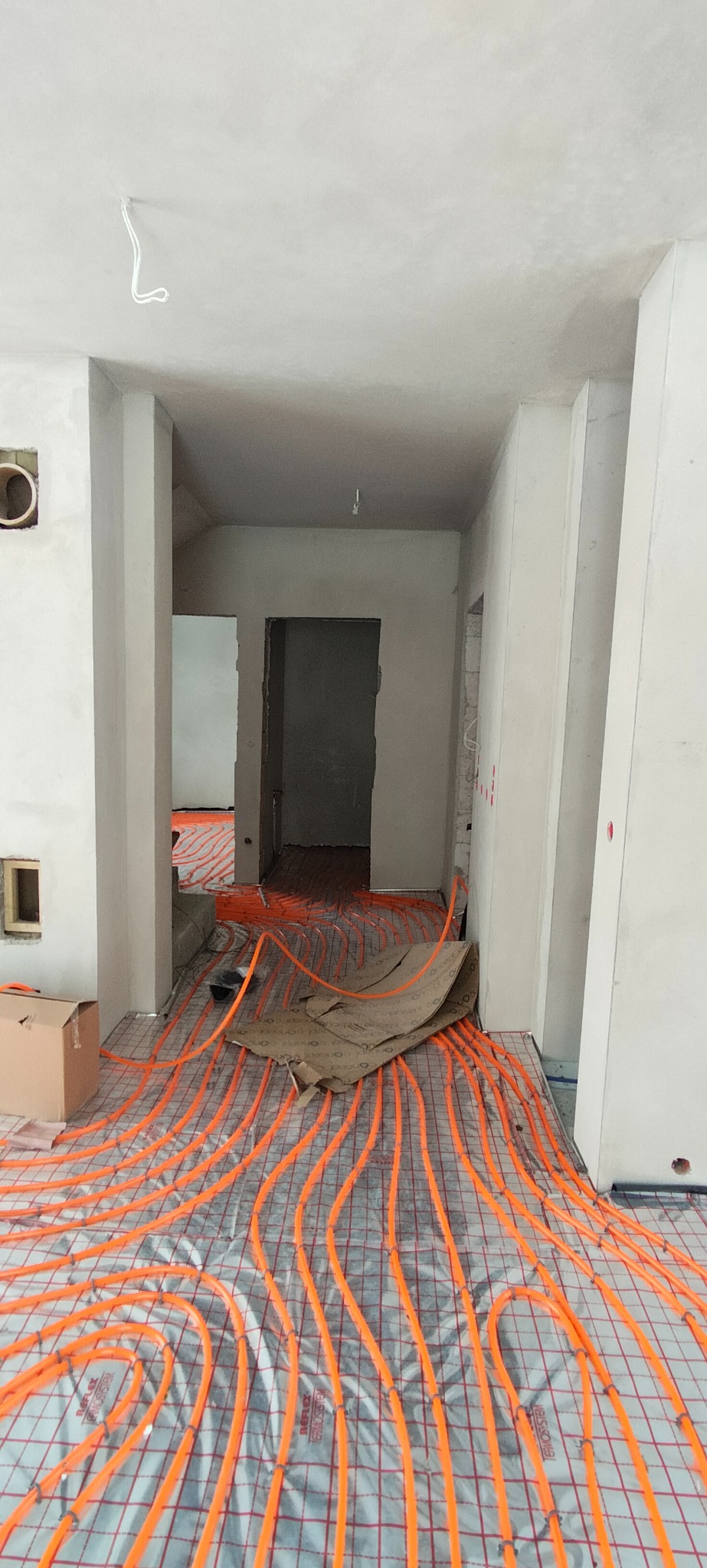 Pomieszczenie w trakcie prac wykończeniowych z widoczną instalacją ogrzewania podłogowego, świeżo położonymi tynkami i przygotowanymi otworami w ścianach.