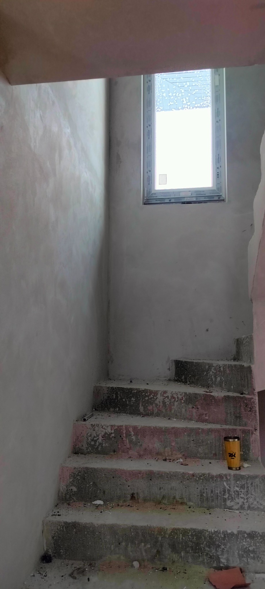 Surowe schody w trakcie prac wykończeniowych, ściany z nałożoną warstwą gładzi, okno w tle. Widoczny fragment puszki z farbą na schodach.