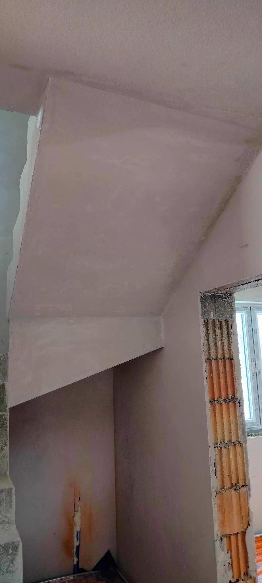 Wnętrze w trakcie prac wykończeniowych: gładkie ściany i sufit w jasnym odcieniu, fragment okna i surowy mur z cegieł. Widoczne ślady po instalacjach.