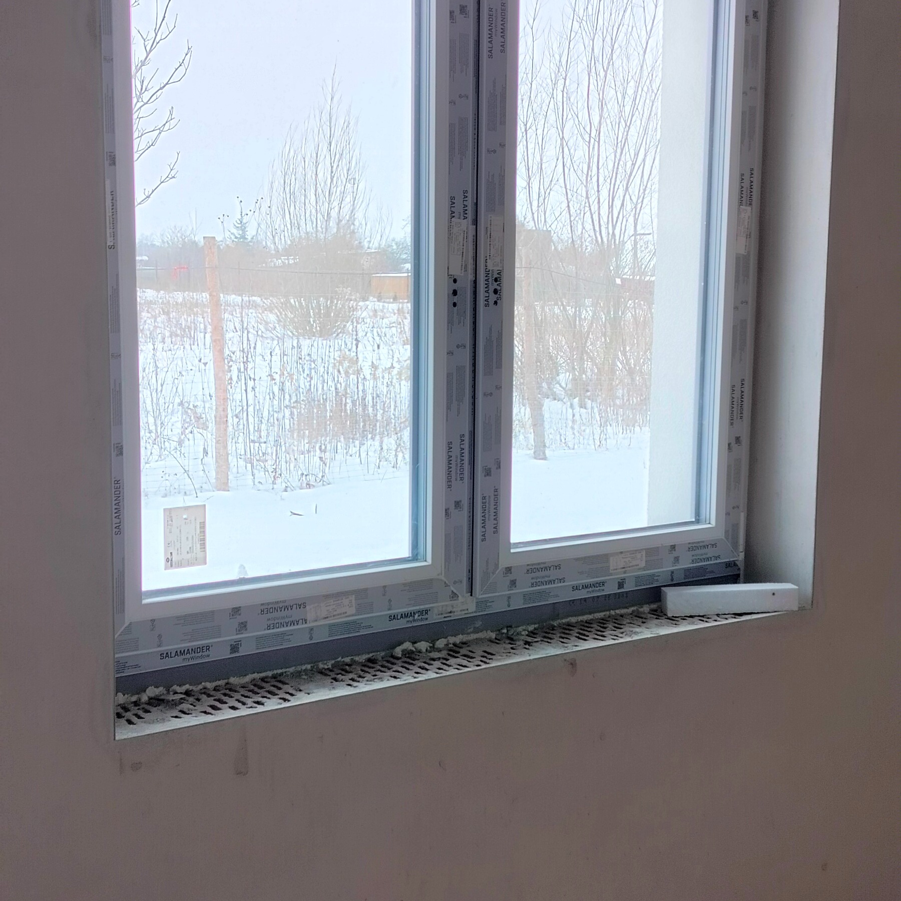 Nowe okno PCV marki Salamander, zamontowane w surowym otworze okiennym. Widoczne uszczelnienia i elementy montażowe. Za oknem zimowy krajobraz.