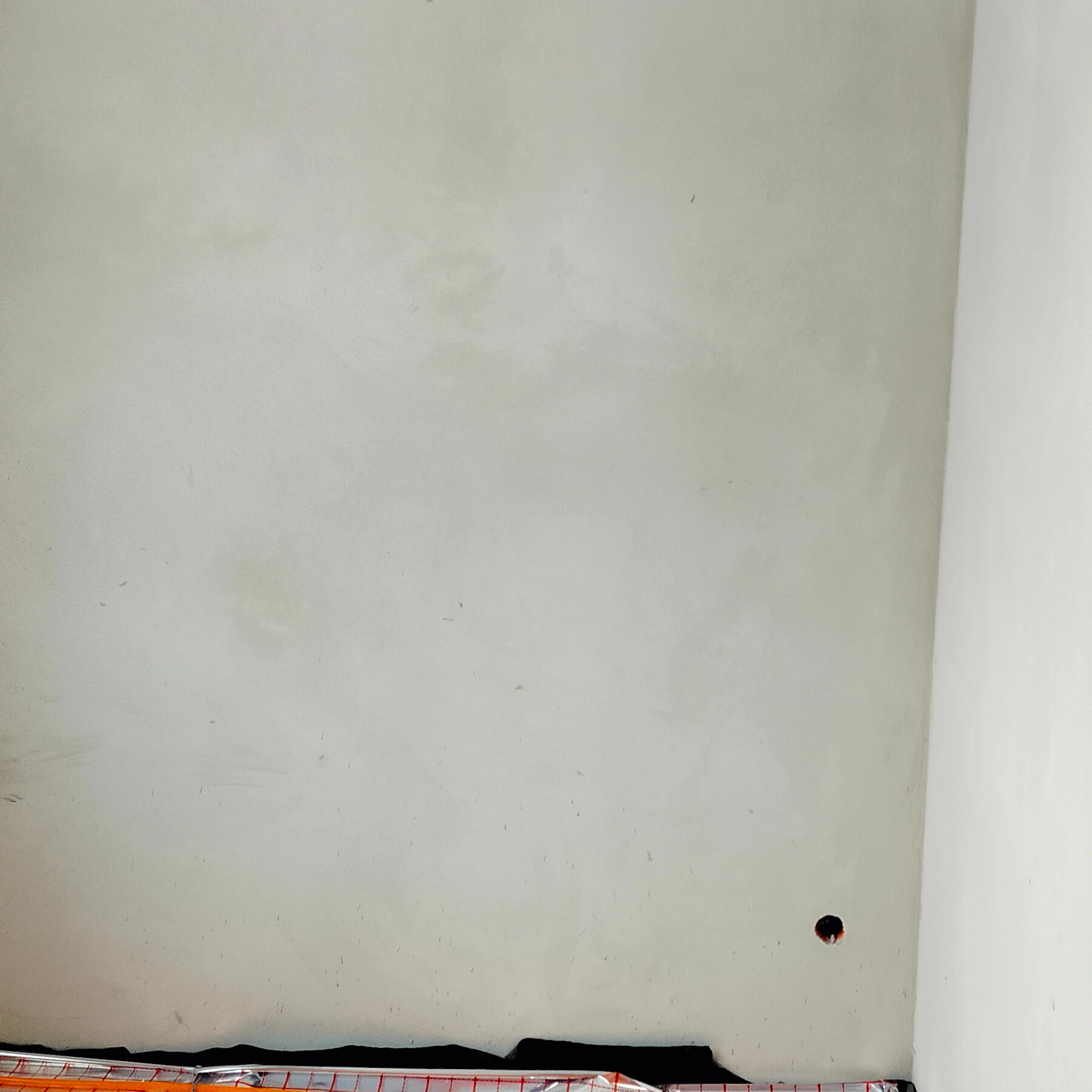 Świeżo położona, jasnoszara gładź na ścianie z widocznym otworem na gniazdko elektryczne i zabezpieczeniem podłogi folią malarską. Minimalistyczne ujęcie.