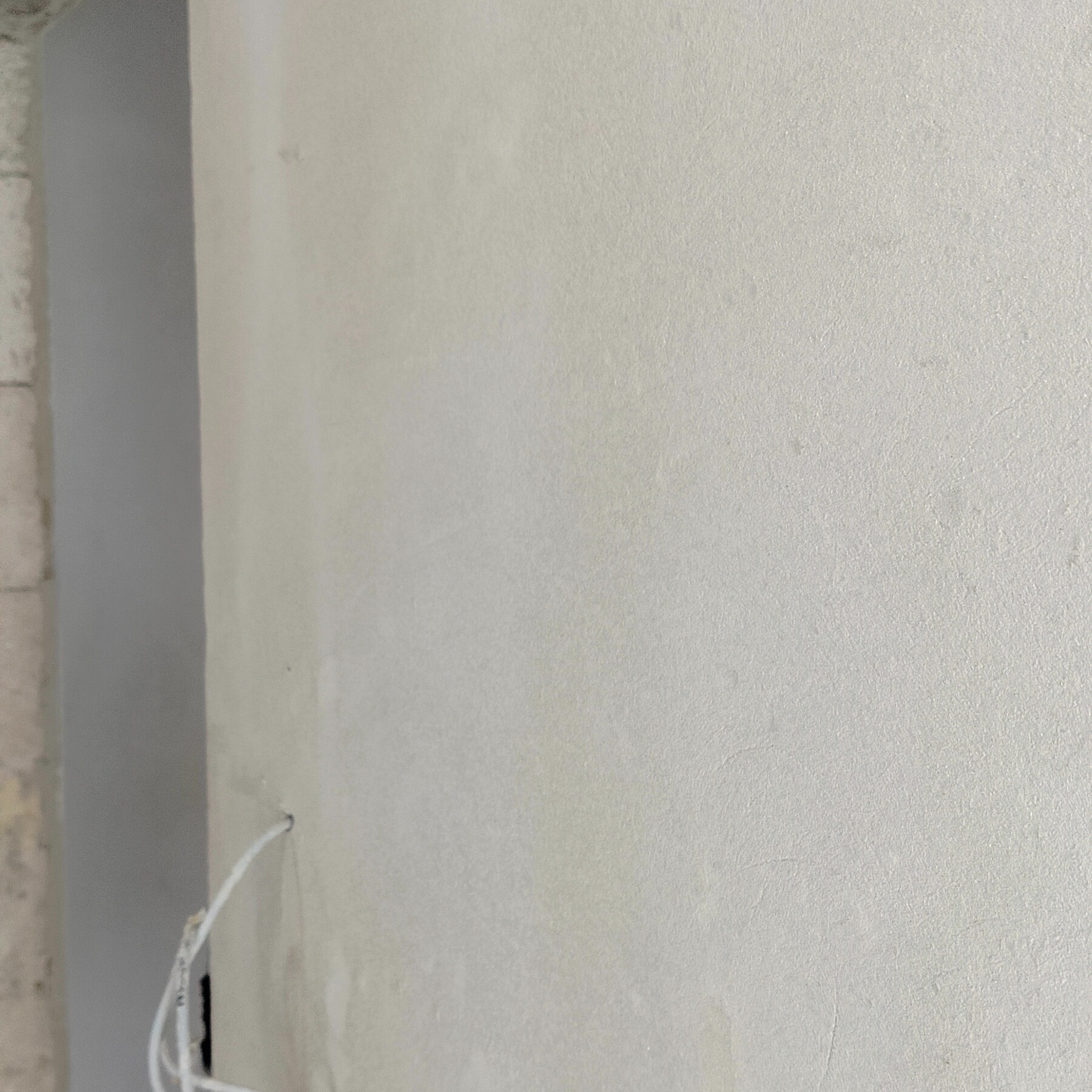 Gładka, jasna ściana po nałożeniu gładzi, z widocznym fragmentem surowego muru i wystającym przewodem elektrycznym. Delikatna faktura powierzchni.