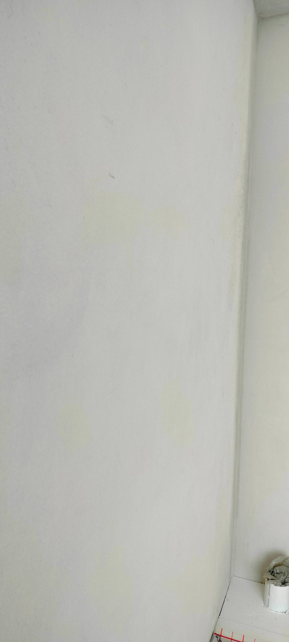 Gładka, biała ściana w rogu pomieszczenia, z delikatnymi smugami i niedoskonałościami, ukazująca efekt końcowy prac wykończeniowych. Widoczne elementy instalacji grzewczej przy podłodze.