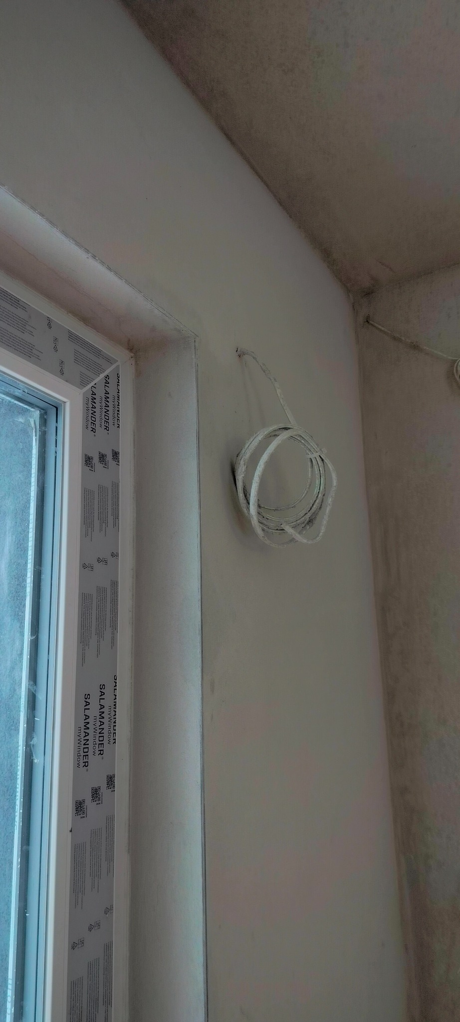 Ściana po nałożeniu gładzi, widoczne przewody elektryczne zwisające ze ściany. Fragment okna z ramą Salamander. Narożnik ściany i sufitu z widocznymi zabrudzeniami.