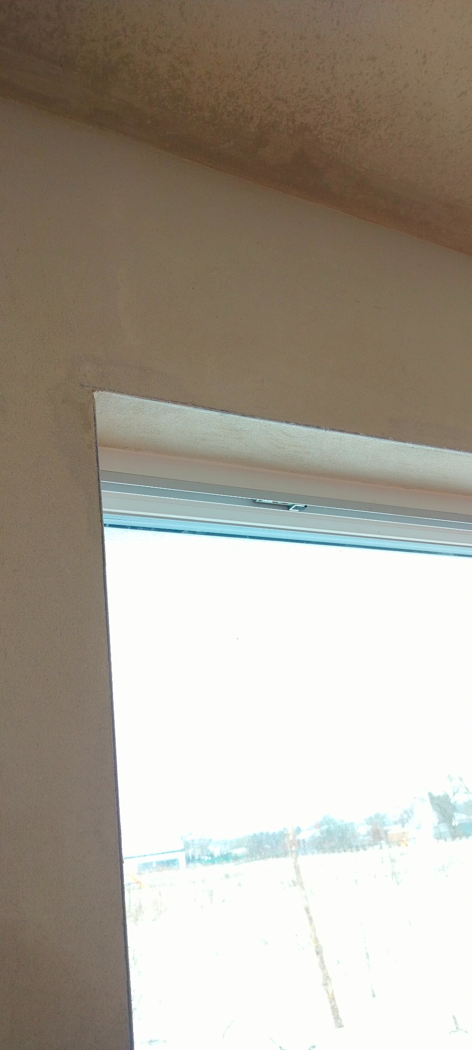 Fragment ściany z oknem po nałożeniu tynku. Widoczne nierówności na suficie i ścianie. Okno białe, plastikowe. Na zewnątrz widoczny zimowy krajobraz.