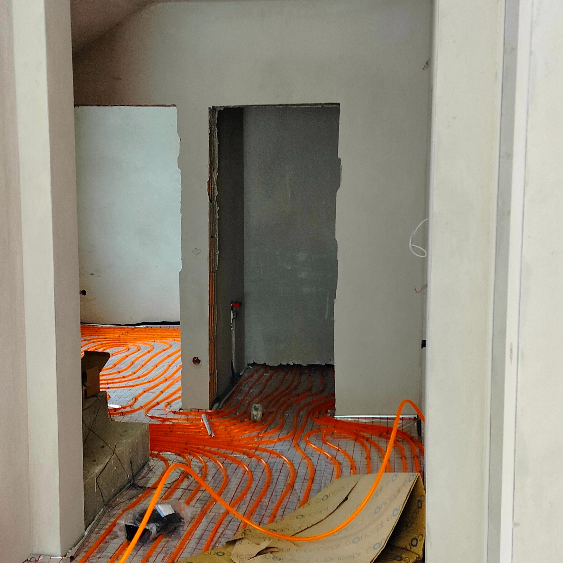 Pomieszczenie w trakcie remontu z widoczną instalacją ogrzewania podłogowego w kolorze pomarańczowym, ułożoną na srebrnej macie. Widoczne surowe ściany i otwory drzwiowe.