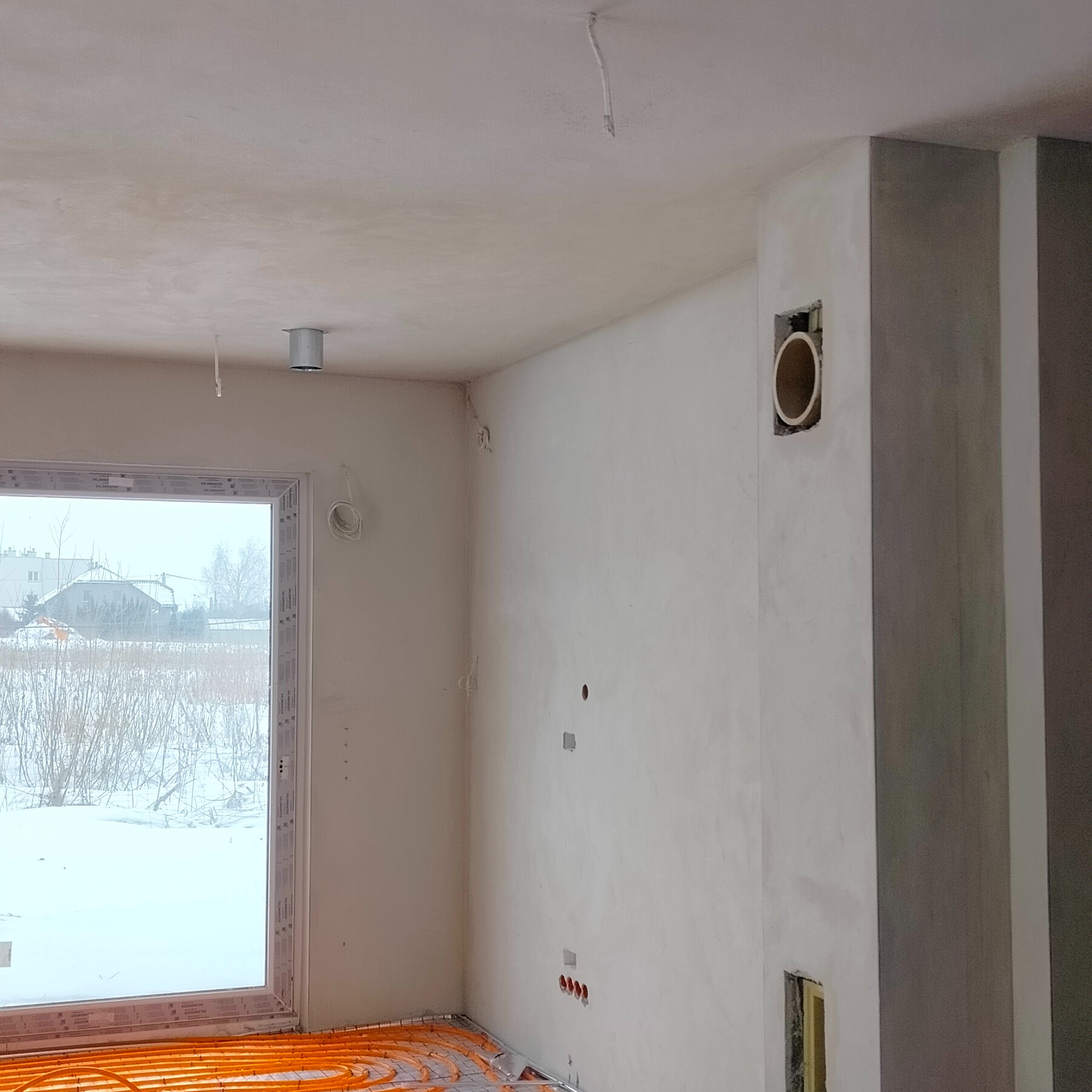 Wnętrze w stanie deweloperskim: ściany po gładziowaniu, instalacja ogrzewania podłogowego, widoczne przewody elektryczne i wentylacyjne. Za oknem zimowy krajobraz.