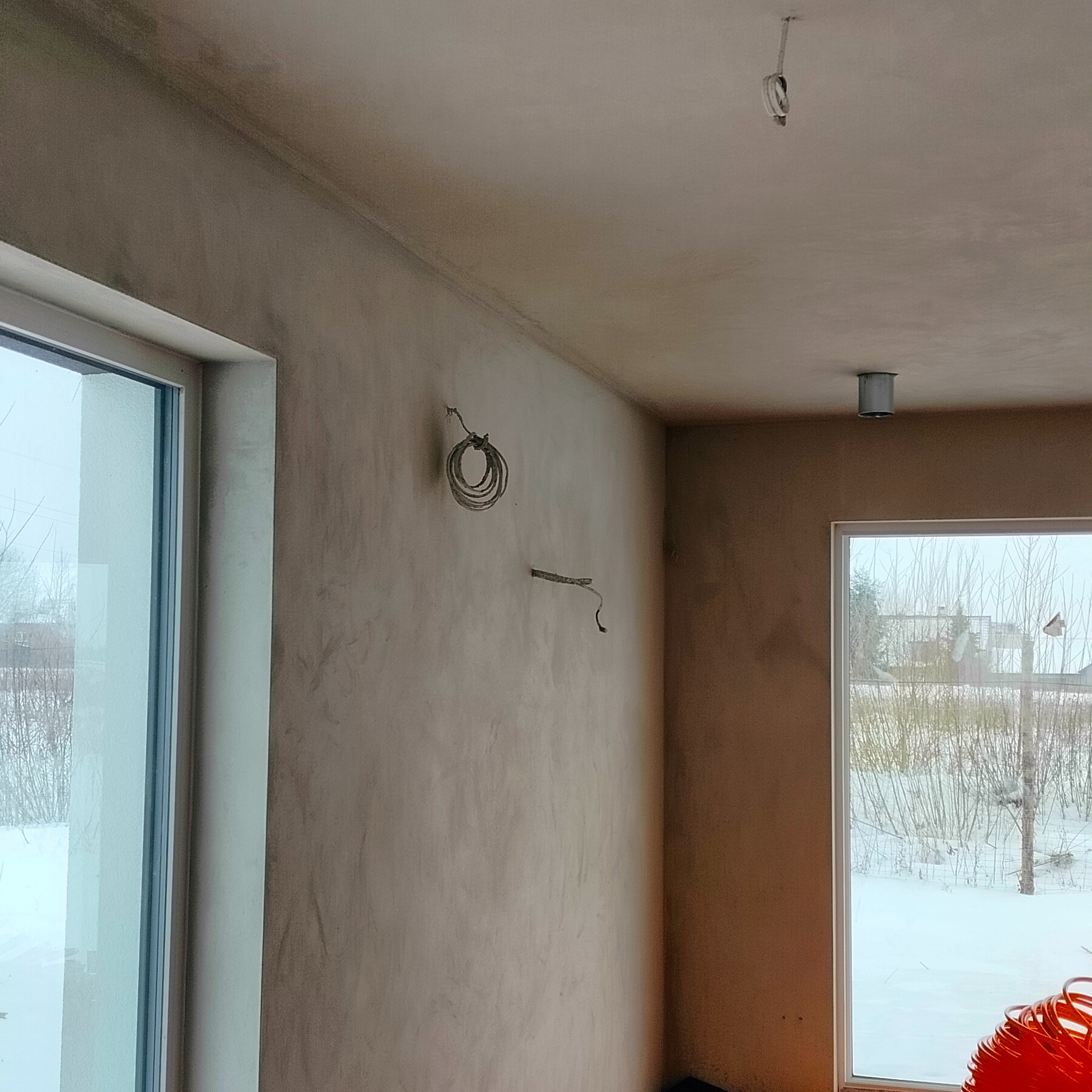 Wnętrze w stanie surowym: ściany pokryte świeżym tynkiem, widoczne przewody elektryczne, okna z widokiem na zimowy krajobraz. Minimalistyczne oświetlenie sufitowe.