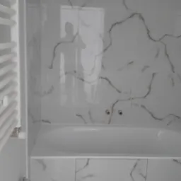 Wykończona łazienka w Kozłowie z wanną obudowaną białymi płytkami imitującymi marmur, z widocznym grzejnikiem drabinkowym po lewej stronie i baterią wannową z czerwonym i niebieskim kurkiem.