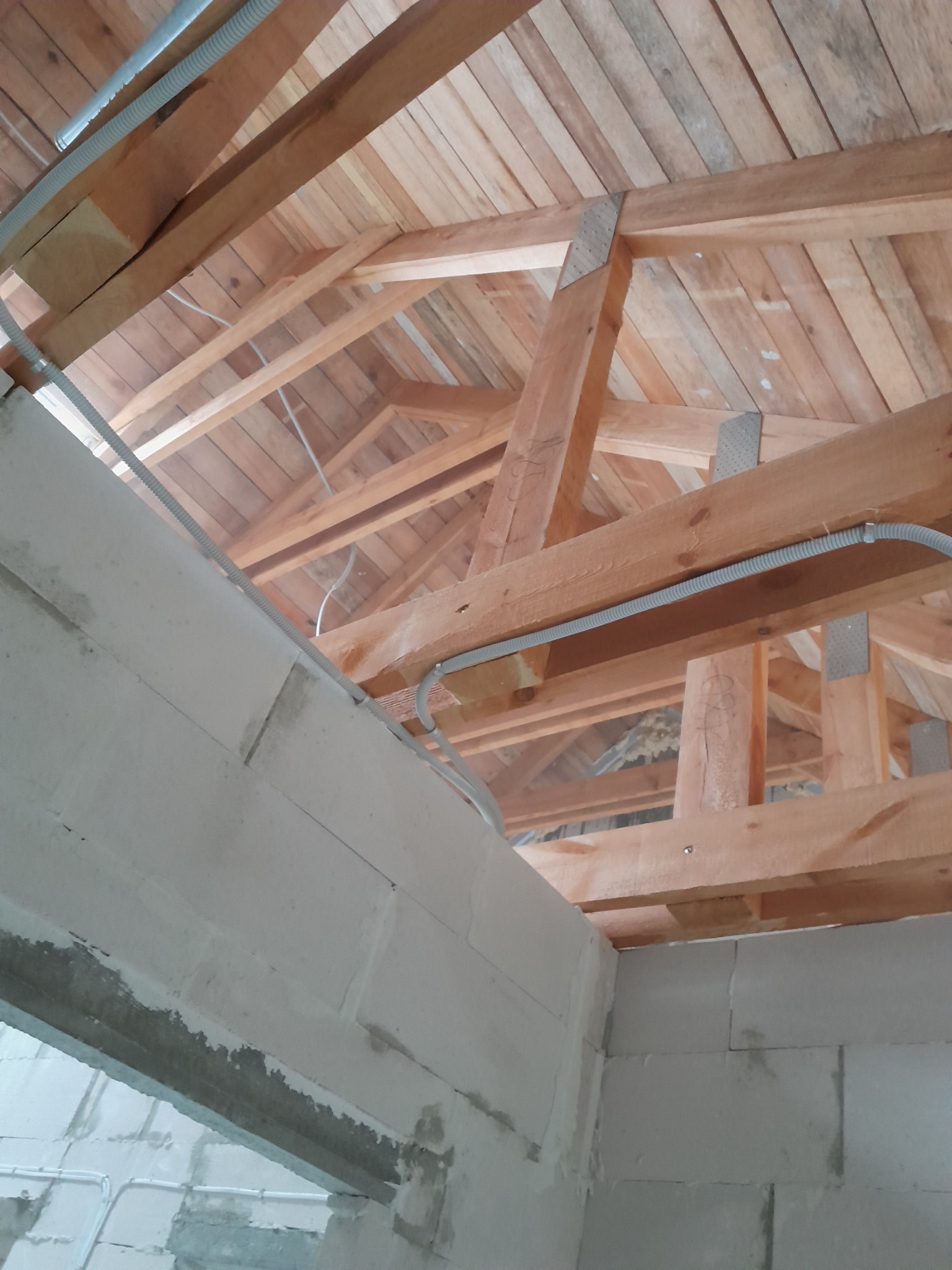 Surowy stan poddasza z drewnianą konstrukcją dachu i fragmentem ściany z betonu komórkowego, widoczne peszle elektryczne.