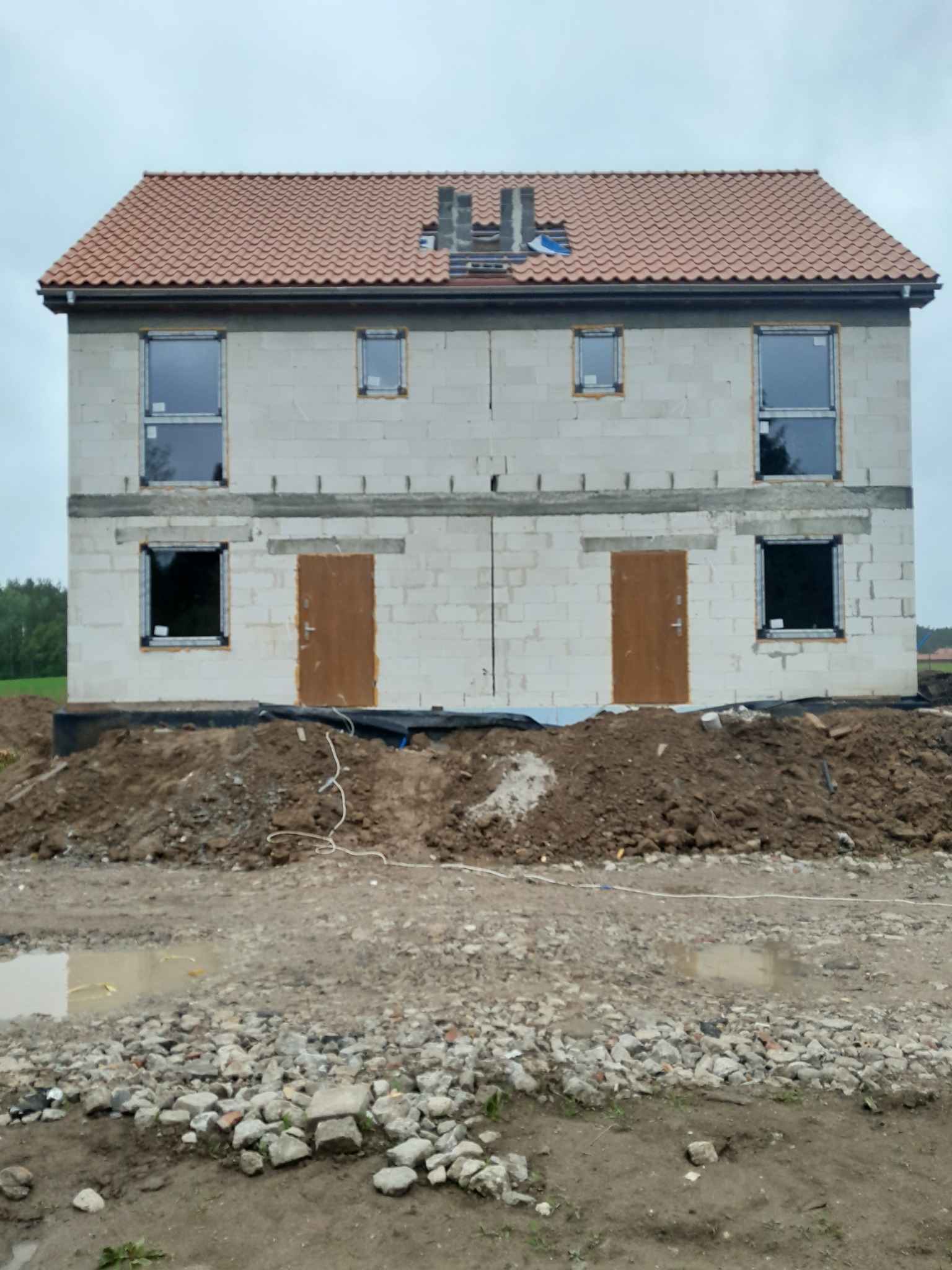 Surowy stan dwukondygnacyjnego budynku z pustaków z częściowo położonym dachem z czerwonej dachówki, widoczne osadzone okna i drzwi, teren wokół budynku nierówny z gruzem i błotem.