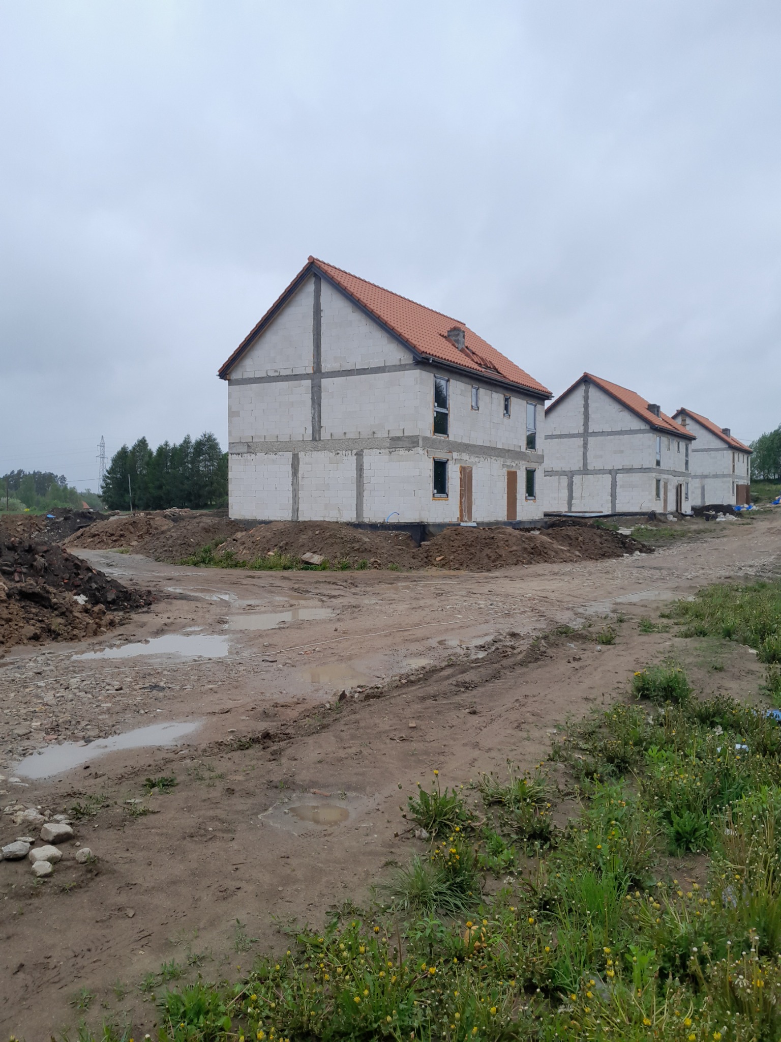 Szereg surowych, dwukondygnacyjnych domów murowanych z czerwonymi dachami w trakcie budowy, widoczne ślady błota i hałdy ziemi na placu budowy, pochmurne niebo.