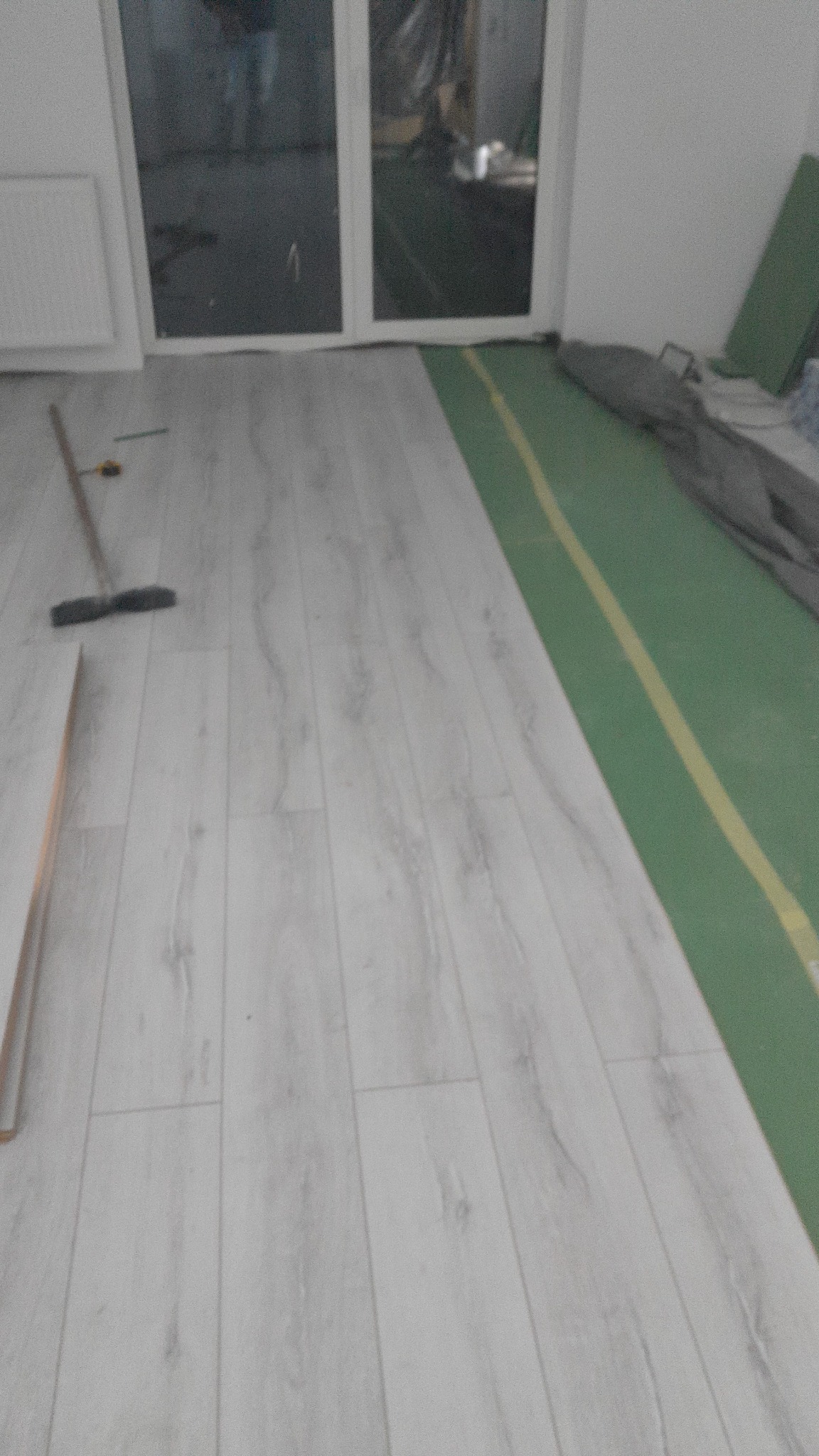 Jasne panele podłogowe ułożone w pokoju z widocznym przygotowaniem do dalszych prac, w tle okno i zielona folia ochronna na podłodze.