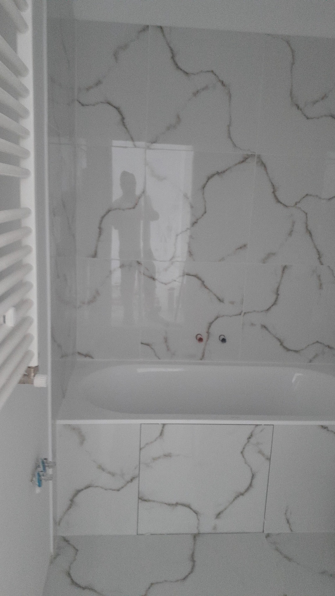 Wykończona łazienka w Kozłowie z wanną obudowaną białymi płytkami imitującymi marmur, z widocznym grzejnikiem drabinkowym po lewej stronie i baterią wannową z czerwonym i niebieskim kurkiem.