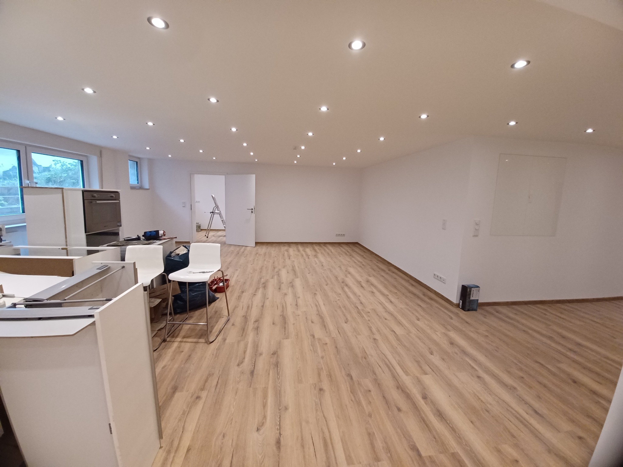 Przestronne wnętrze w trakcie remontu: jasne ściany, podłoga z paneli, częściowo zmontowane meble kuchenne i krzesła barowe, widoczne instalacje elektryczne na suficie.