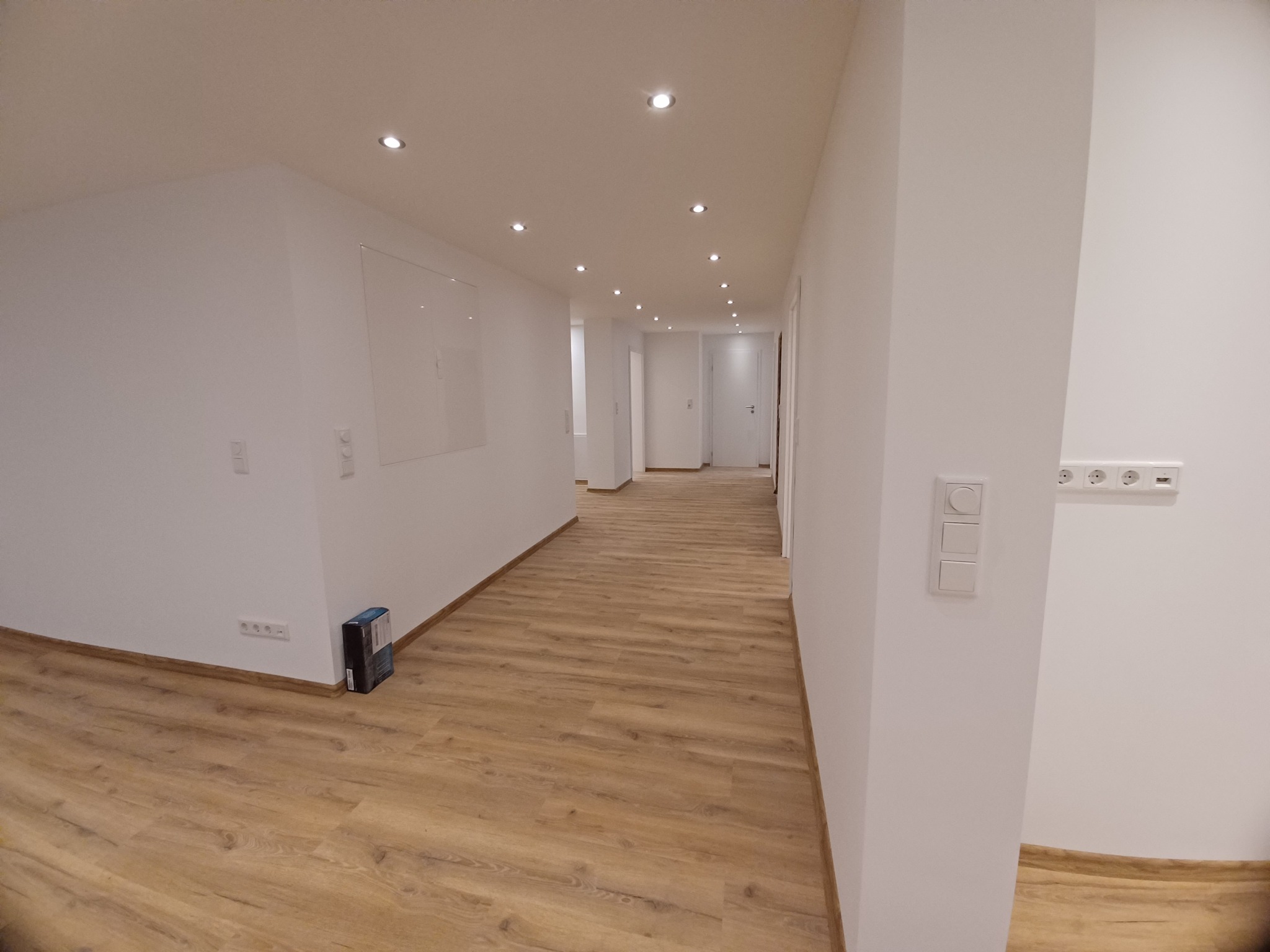 Wnętrze wykończonego korytarza z panelami podłogowymi w odcieniach brązu, białymi ścianami z włącznikami i gniazdkami, oraz punktowym oświetleniem sufitowym, widok w głąb mieszkania.