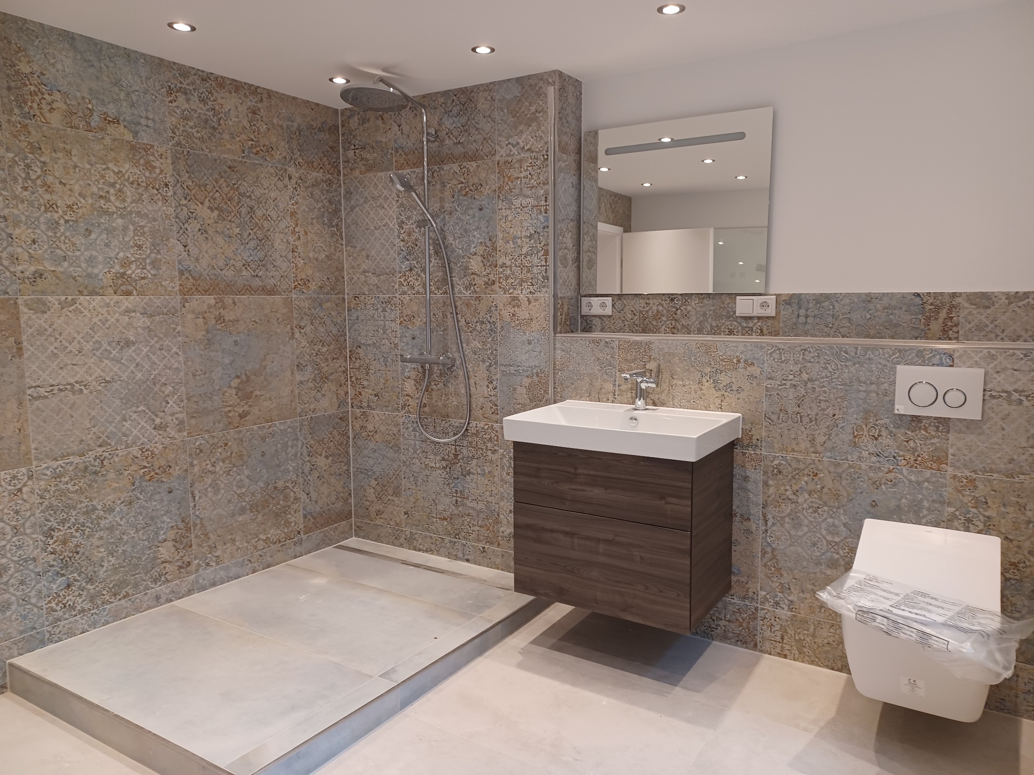 Nowoczesna łazienka z prysznicem typu walk-in, umywalką z szafką w kolorze ciemnego drewna i toaletą w folii ochronnej, wykończona płytkami w stylu vintage.