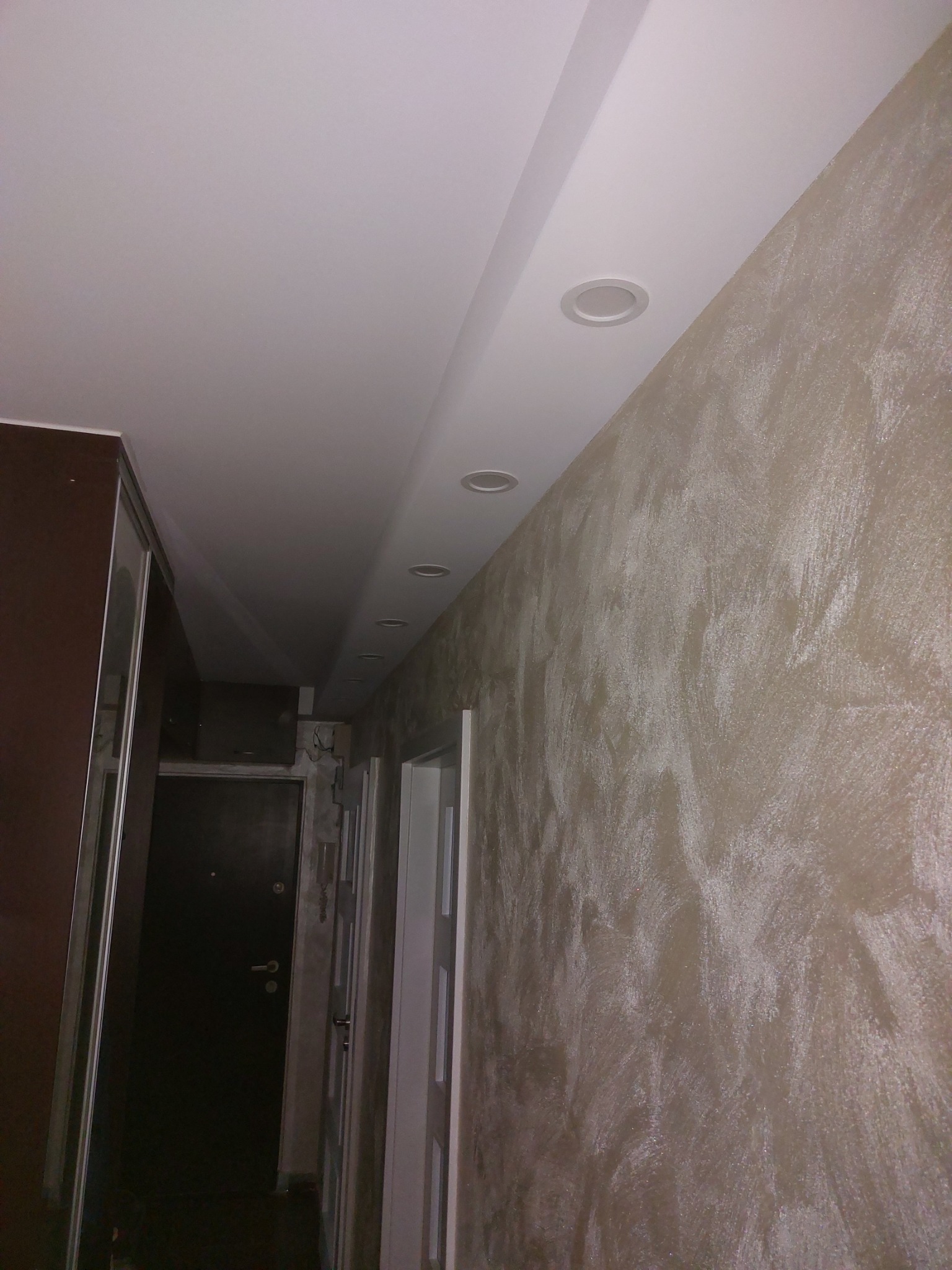 Korytarz po remoncie: ściana pokryta dekoracyjną, beżową strukturą, podwieszany sufit z zamontowanymi, okrągłymi lampami LED, widoczna szafa przesuwna i drzwi wejściowe.