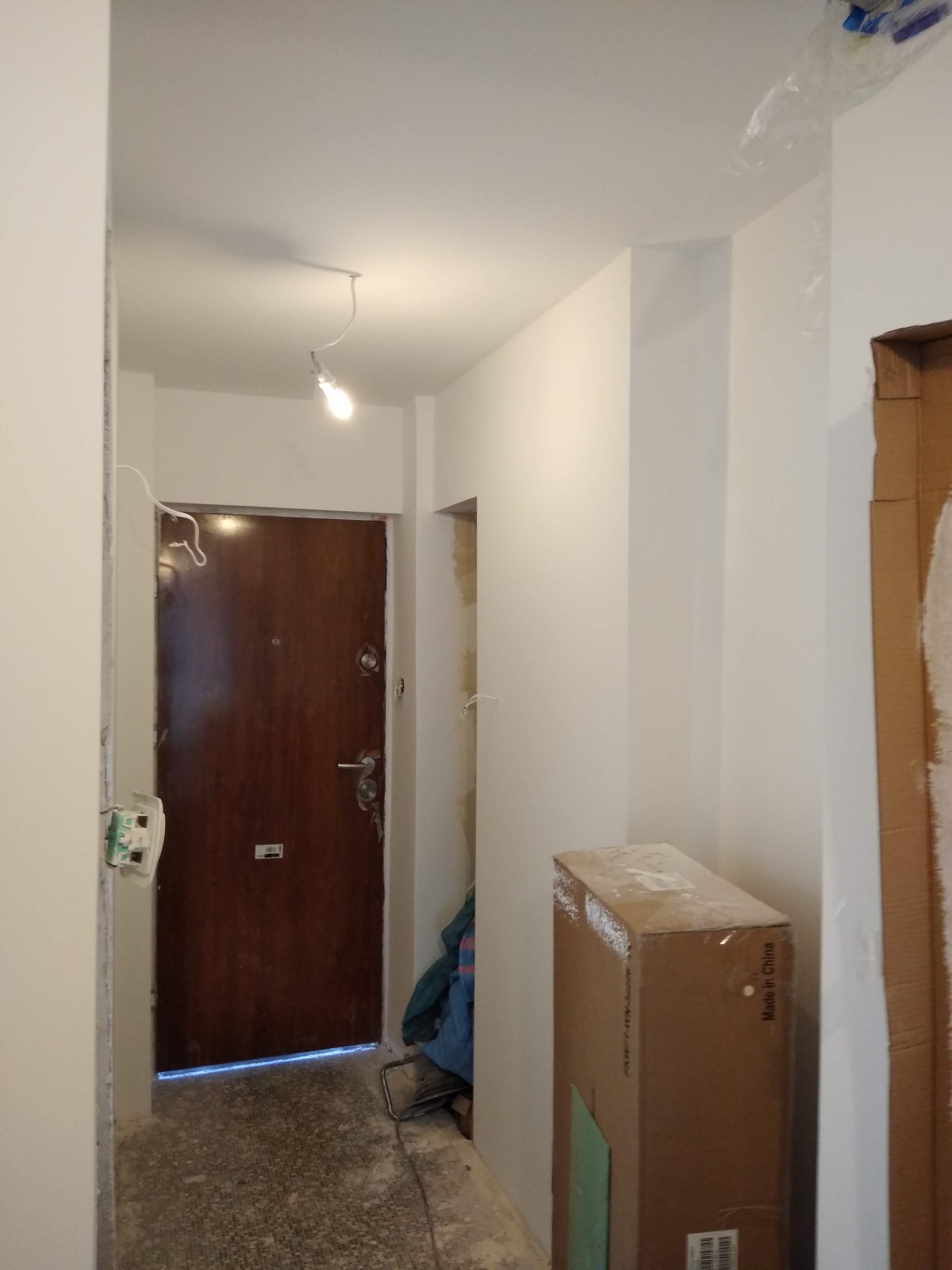 Korytarz w trakcie remontu: świeżo pomalowane ściany na biało, surowy beton na podłodze, kartonowe pudło i zabezpieczona folią framuga drzwi.