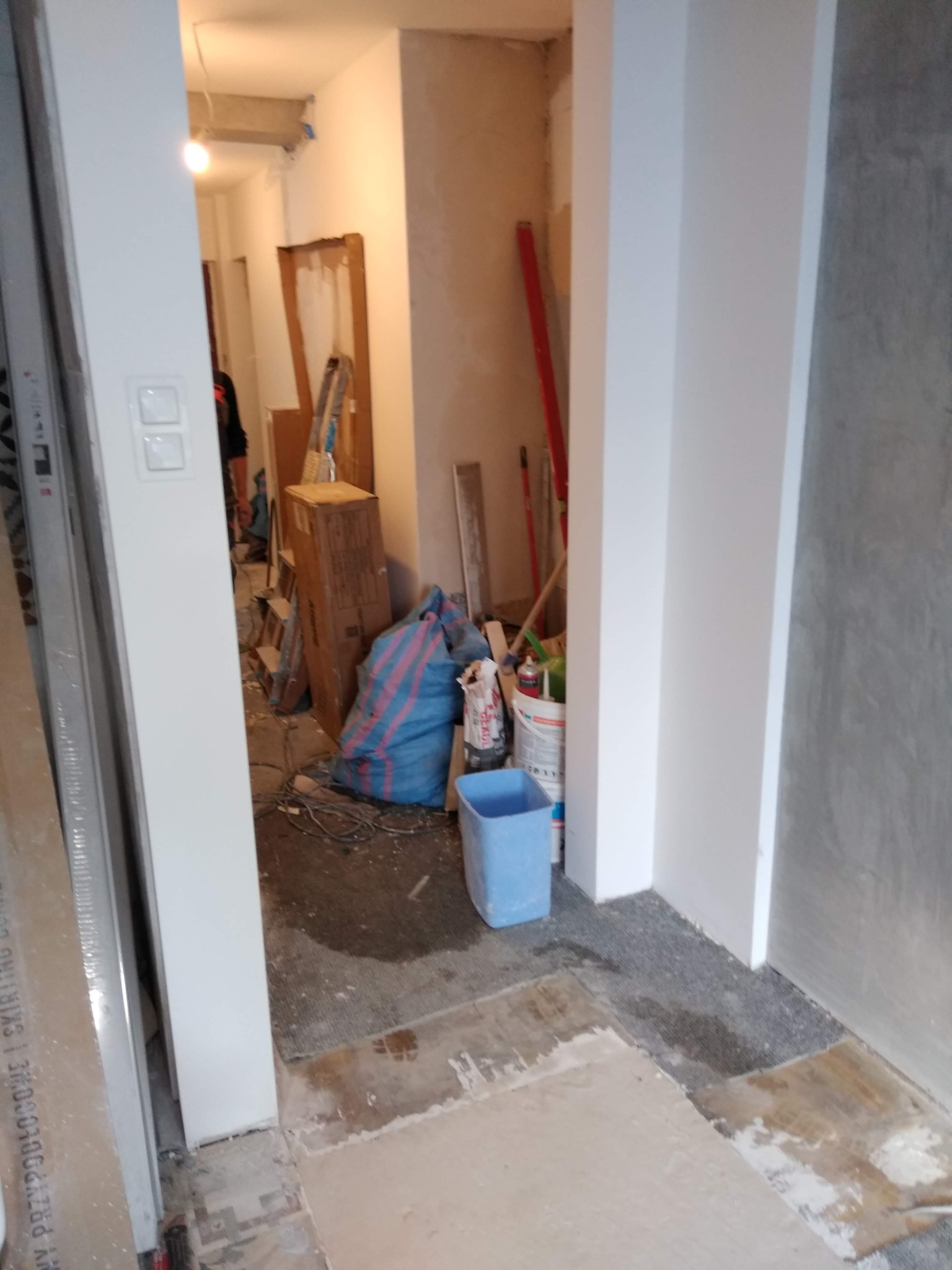 Wnętrze w trakcie remontu: widok na korytarz z workami, wiadrami i narzędziami budowlanymi, surowa podłoga i ściany w trakcie prac wykończeniowych.