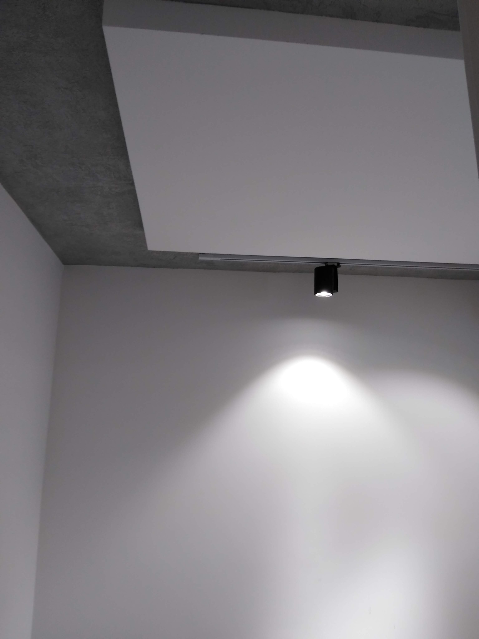 Minimalistyczne wnętrze w Warszawie z gładkimi, białymi ścianami oświetlonymi pojedynczym, punktowym reflektorem na szynie, sufit częściowo wykończony w surowym betonie.