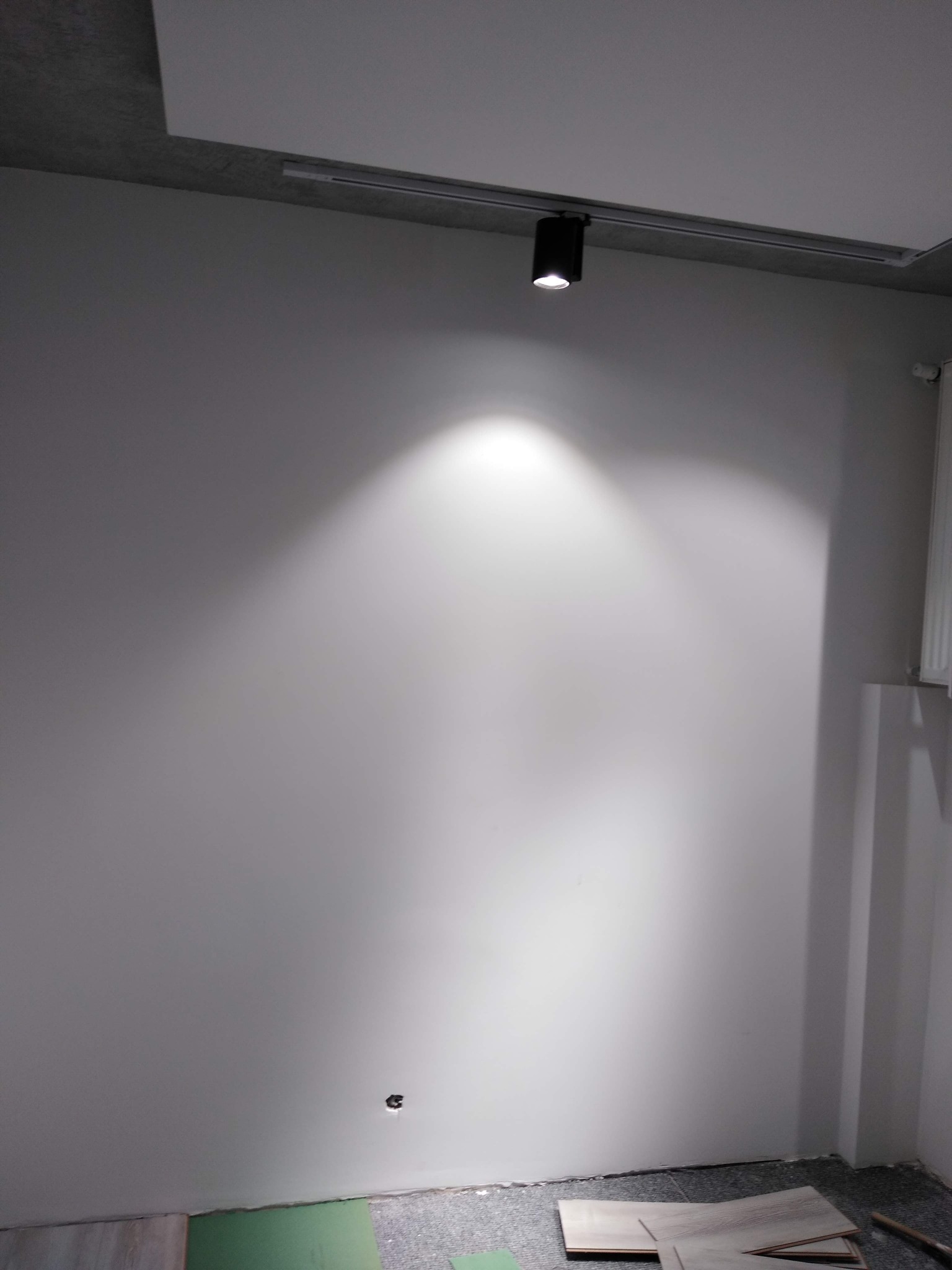 Świeżo pomalowana szara ściana oświetlona reflektorem sufitowym, widoczne fragmenty podłogi z płytami gipsowo-kartonowymi i narzędziem.