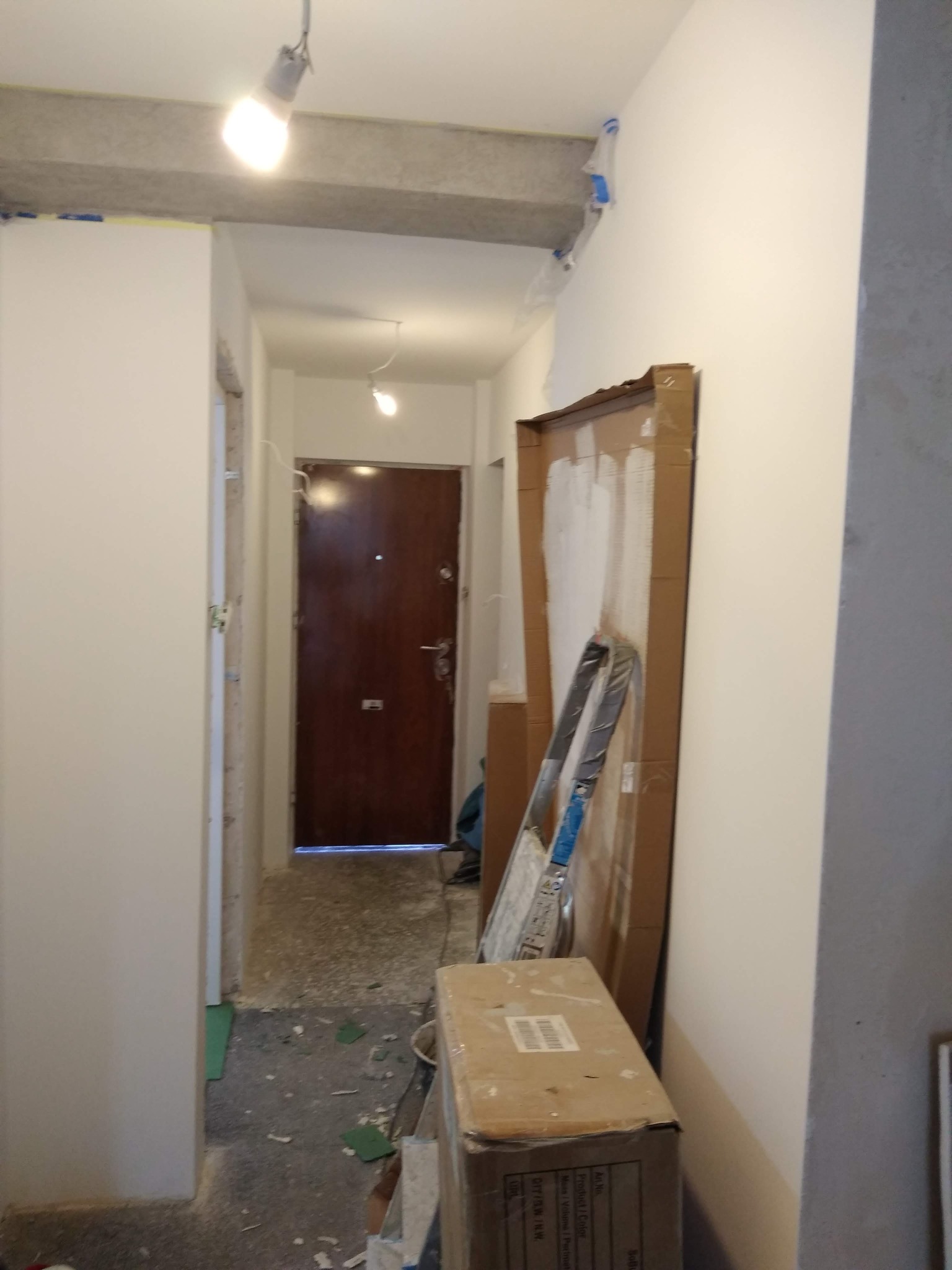 Korytarz w trakcie remontu: widoczne świeżo pomalowane ściany, zabezpieczona folią ościeżnica, kartonowe osłony, drabina oparta o ścianę i karton z materiałami budowlanymi.